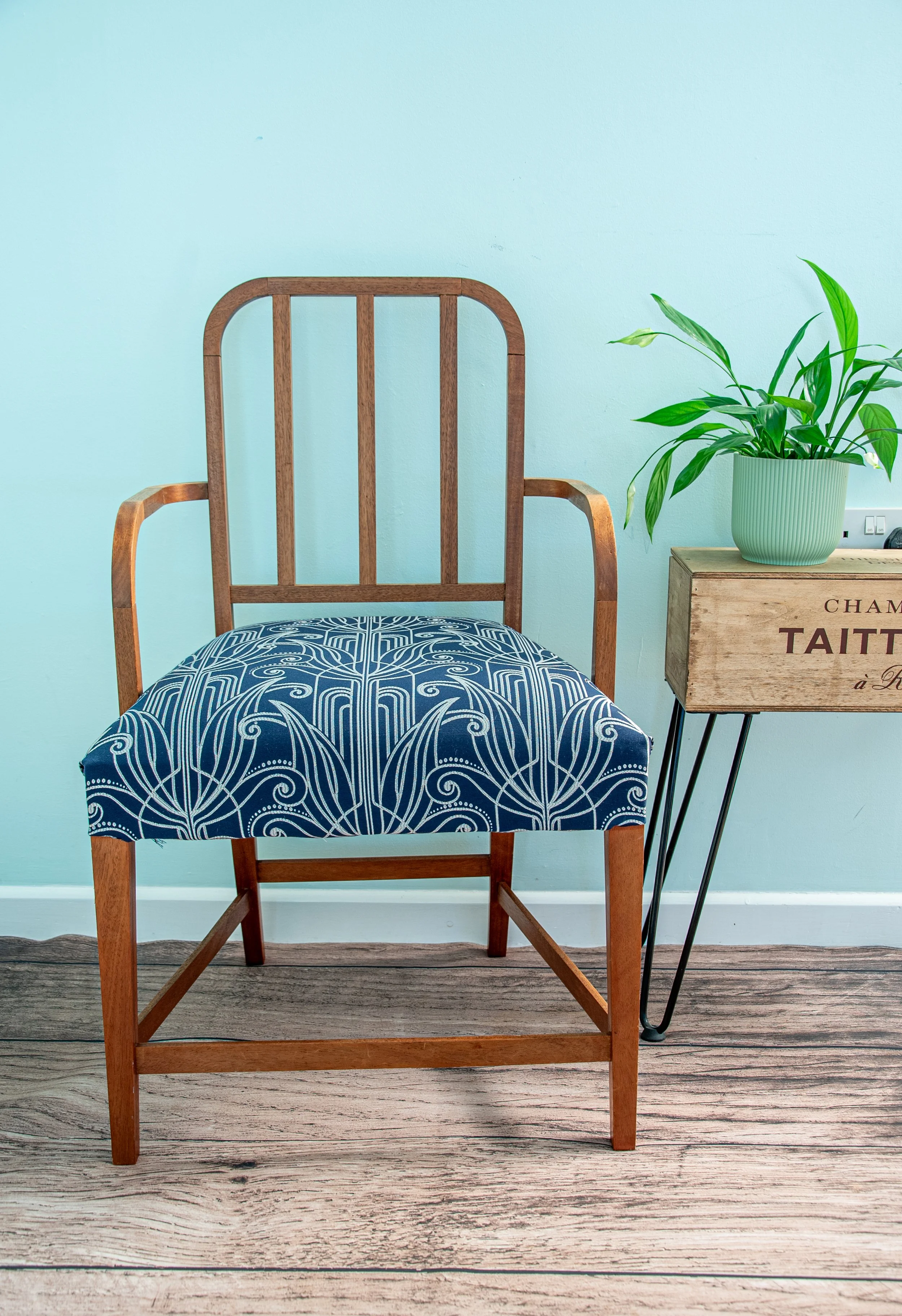 Wooden chair with blue patterned cushion next to a wooden side table with a green potted plant on top, against a light blue wall.
