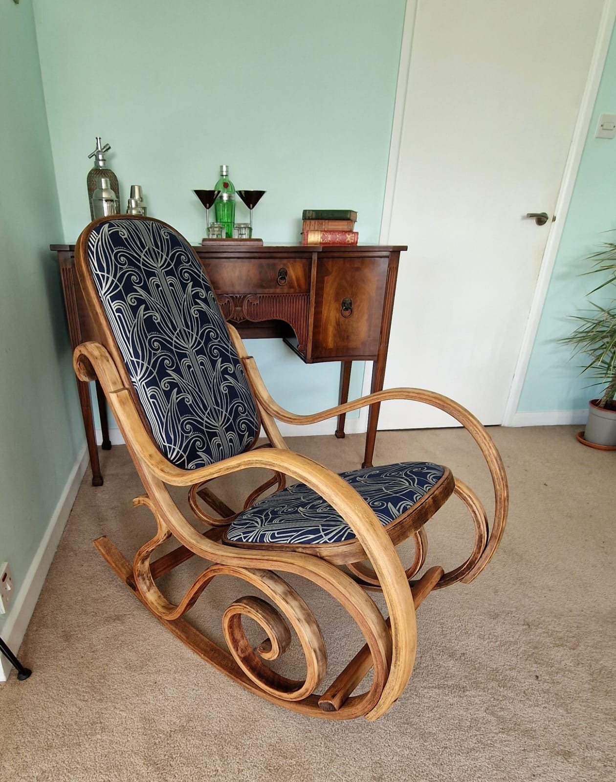 A wooden rocking chair with black and white patterned cushions in front of a vintage wooden sideboard. The sideboard has bottles, glasses, and books on top. A potted plant is nearby, and a white door is in the background.