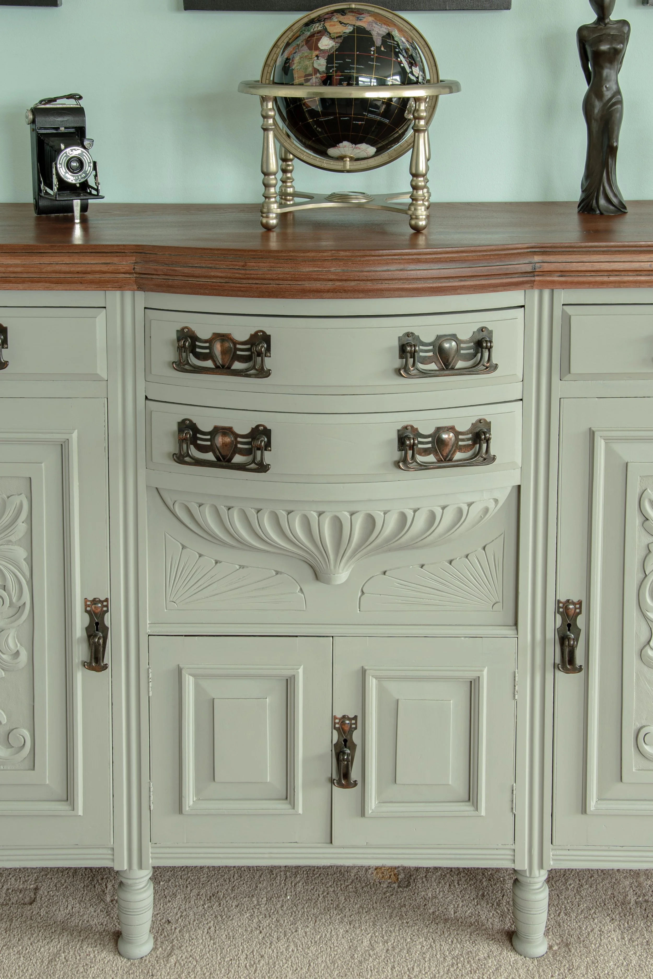 A vintage-style cream-colored sideboard with decorative drawers and cabinet doors, topped with a wooden surface. On the surface, there is a globe on a stand, a small vintage camera, and a dark figurine of a woman.