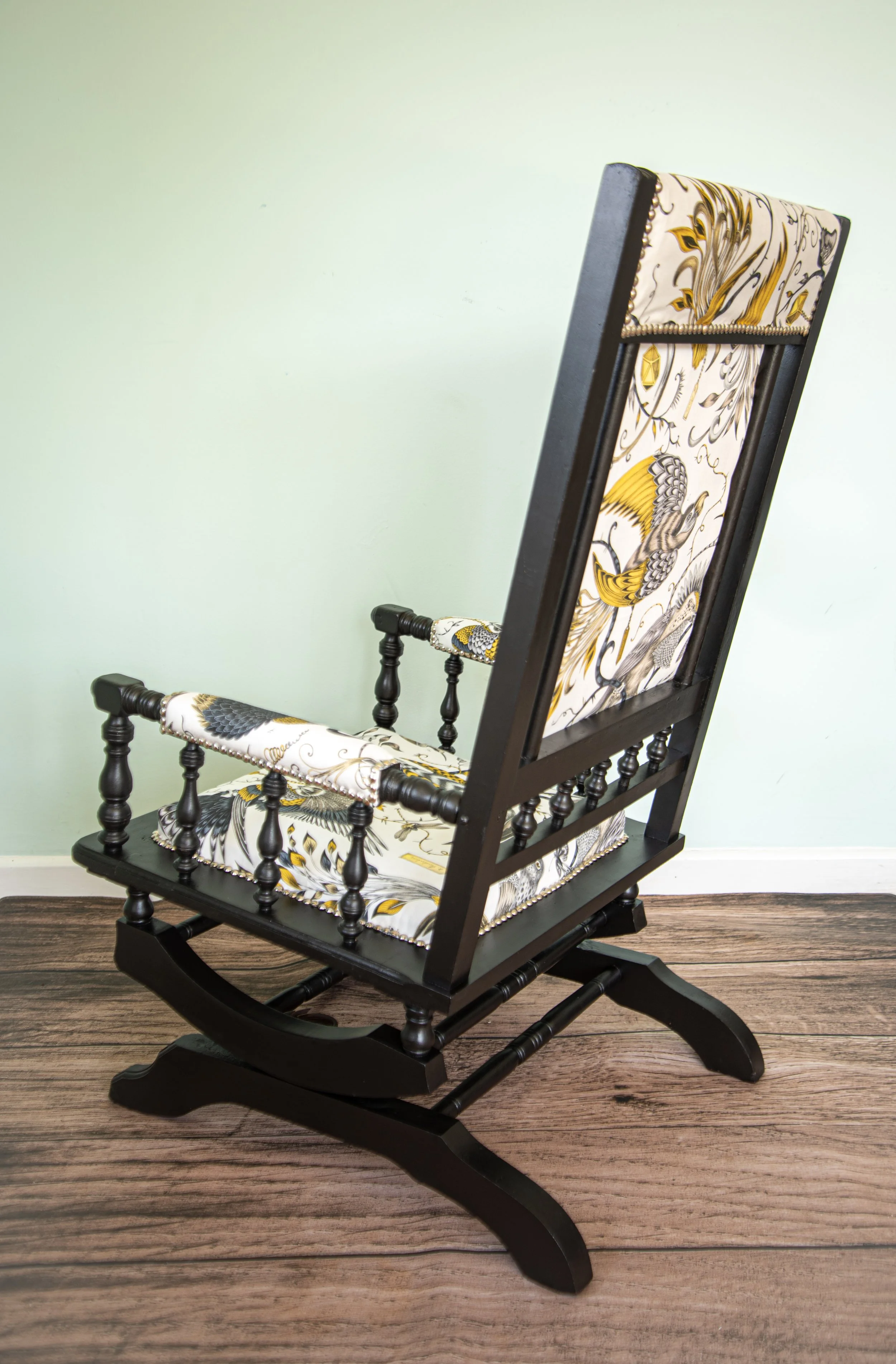 A traditional wooden rocking chair with a tall backrest, upholstered in fabric featuring gold, black, and gray bird and floral patterns, placed on a wooden floor against a plain, light-colored wall.