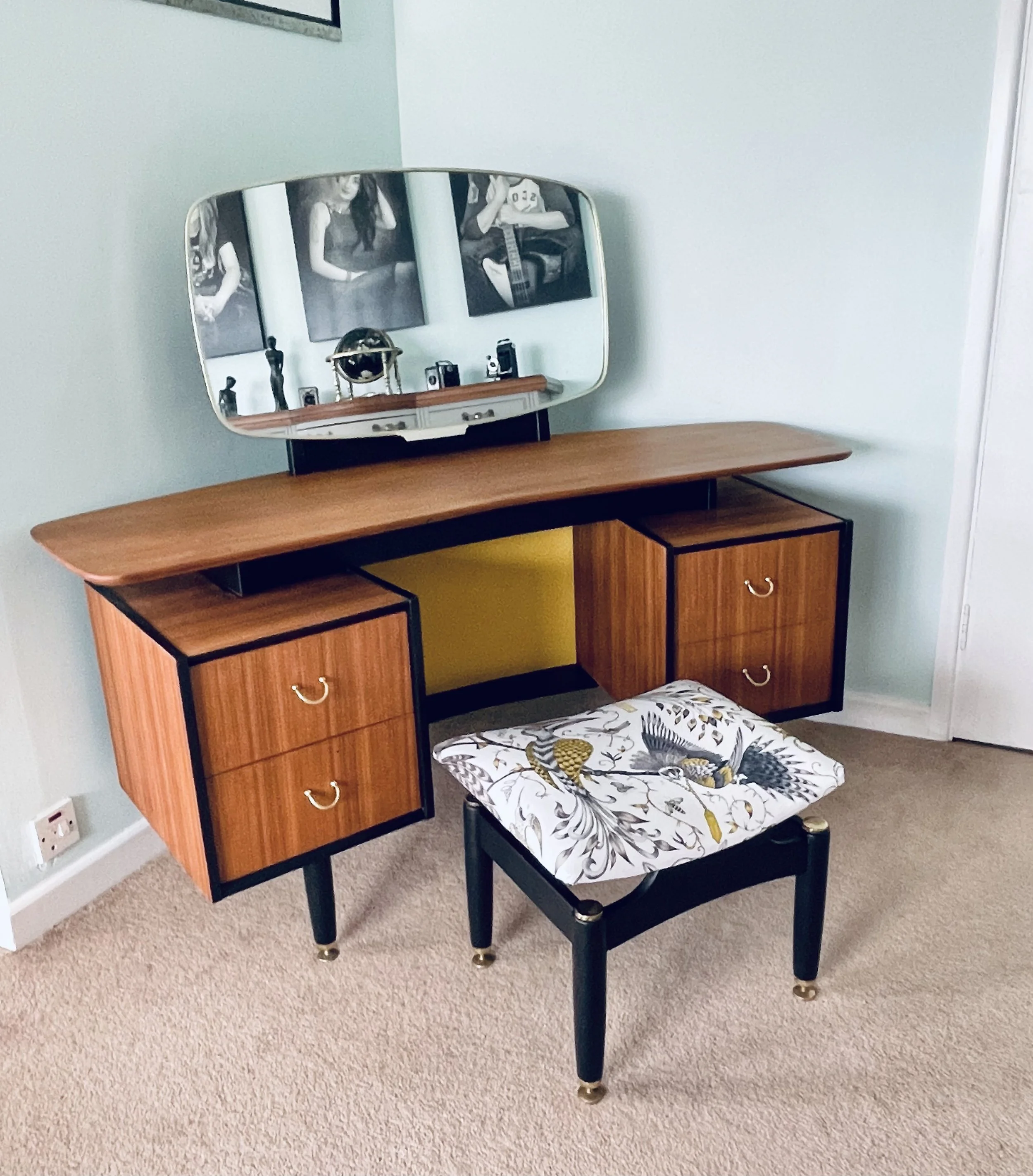 A vintage wooden vanity table with a large oval mirror, decorative drawer handles, and a small cushioned stool with a floral fabric cover, set on a beige carpeted floor in a light green room.