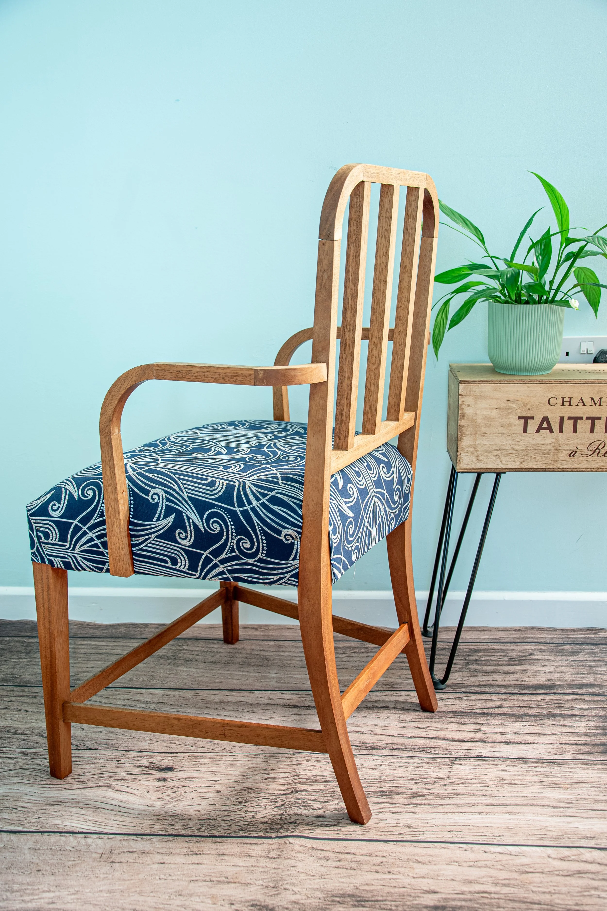 Wooden chair with a blue and white patterned cushion, next to a small wooden side table with a green potted plant on it, against a light blue wall.
