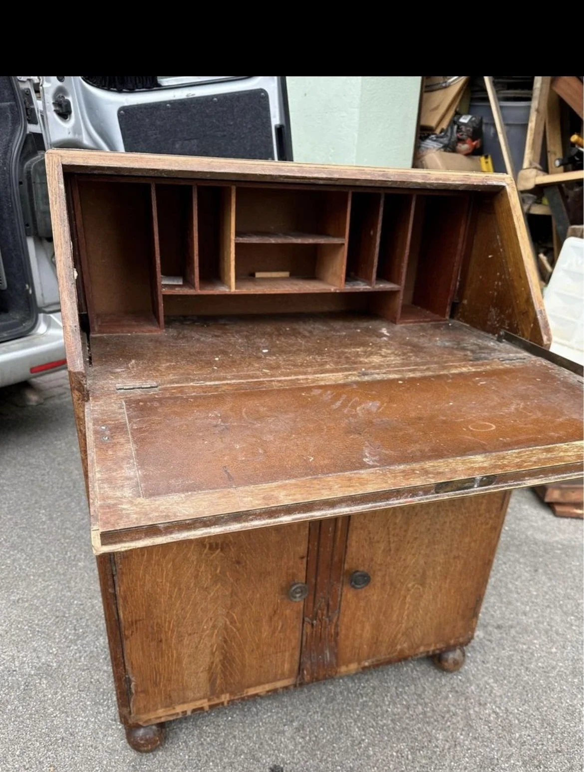 Old wooden desk with a hutch, scratches, and dirt, sitting outdoors near a vehicle and workshop tools.