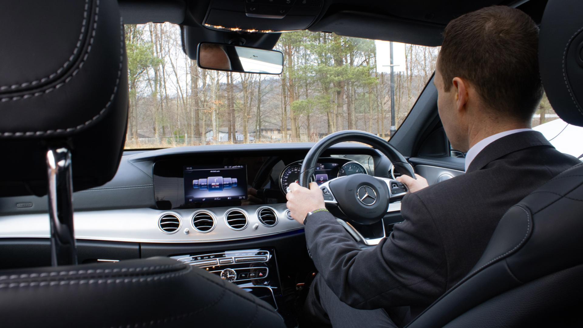 The interior of a luxury Mercedes-Benz car with a man in a suit driving on a road with trees outside.