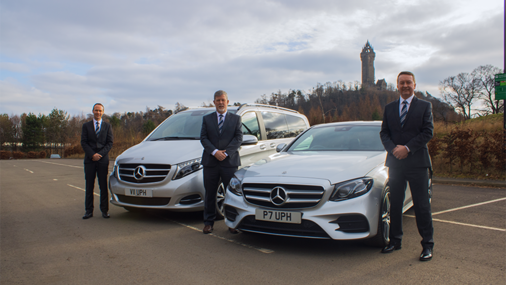 Three men in formal suits standing near silver luxury Mercedes-Benz vehicles in an open parking lot with a historic tower and trees in the background during daytime.