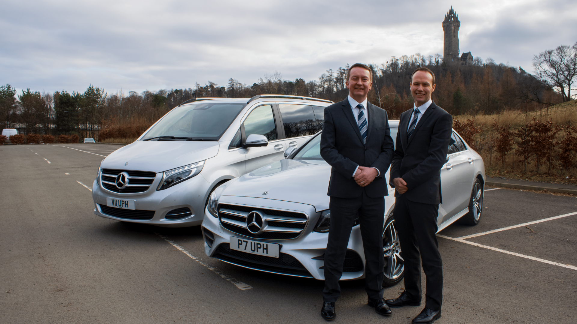 Two men in suits standing in front of parked silver Mercedes-Benz cars, with a historic tower in the background, on a cloudy day in a parking lot.