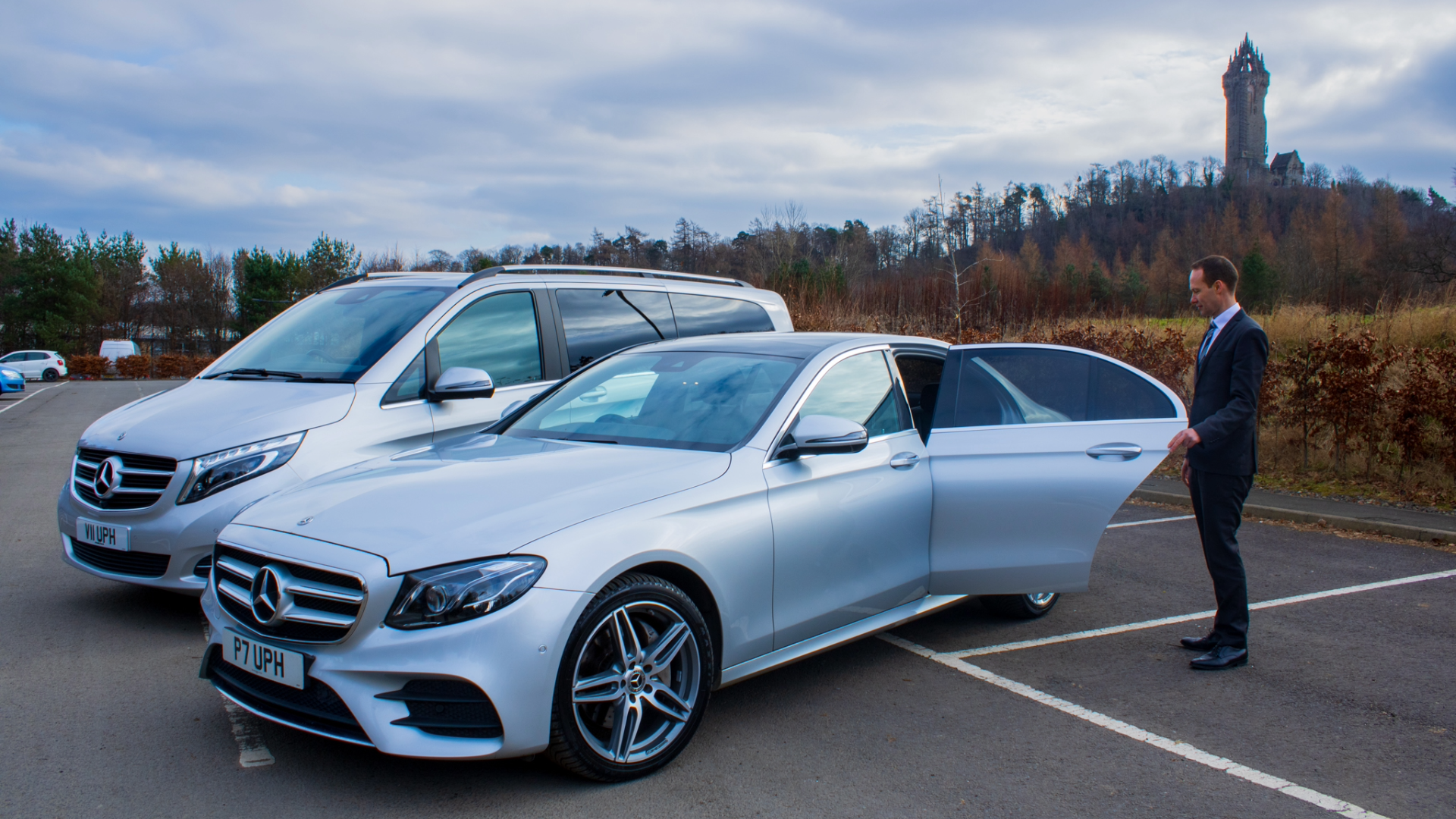 A man in a business suit opening the door of a silver Mercedes-Benz car in a parking lot, with a white van parked next to it and a scenic background including trees and a tall historic tower on a cloudy day.