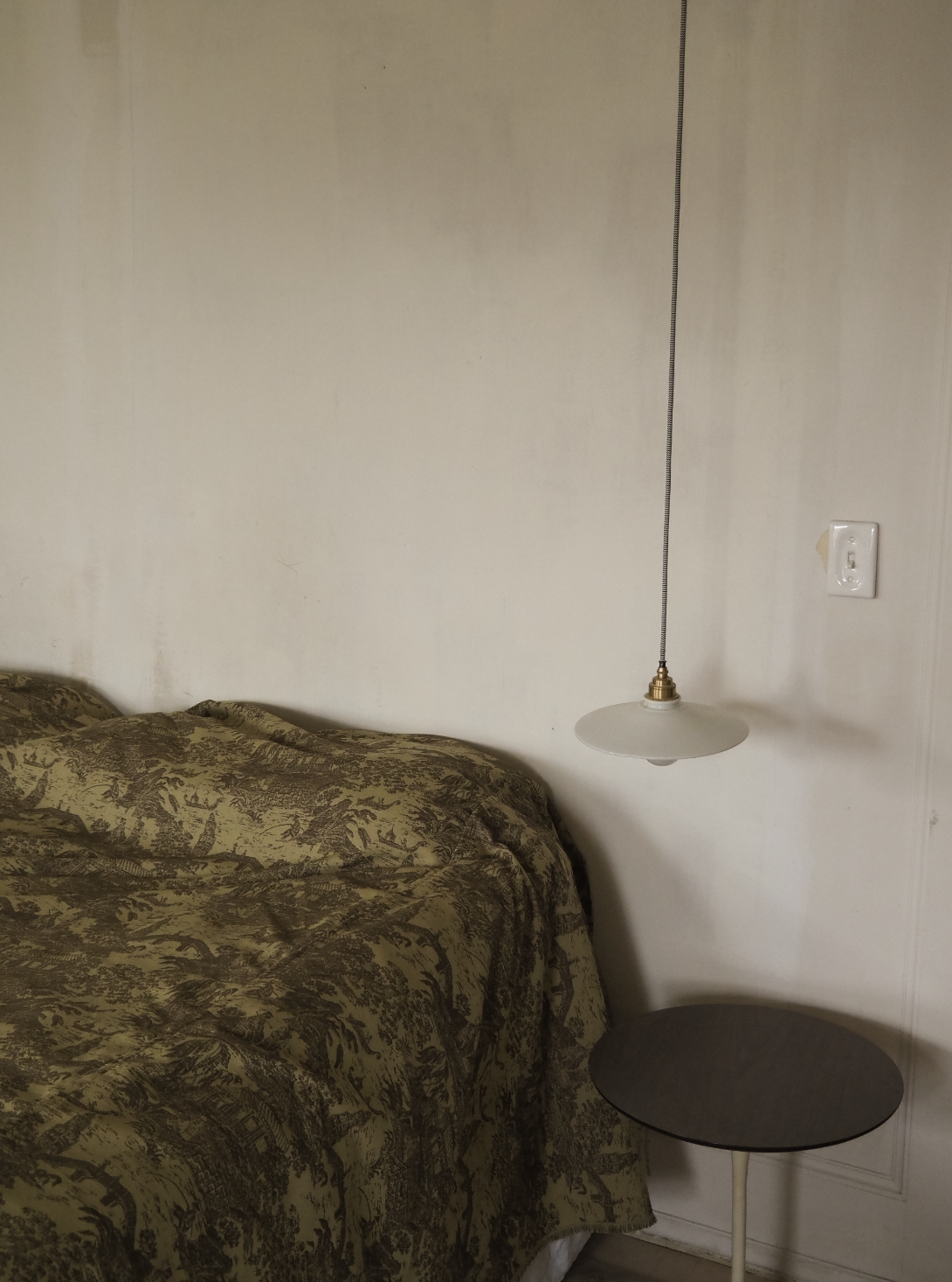 A bedroom corner with a bed covered in a patterned bedspread, a wall-mounted hanging lamp with a pull chain, and a small round side table.