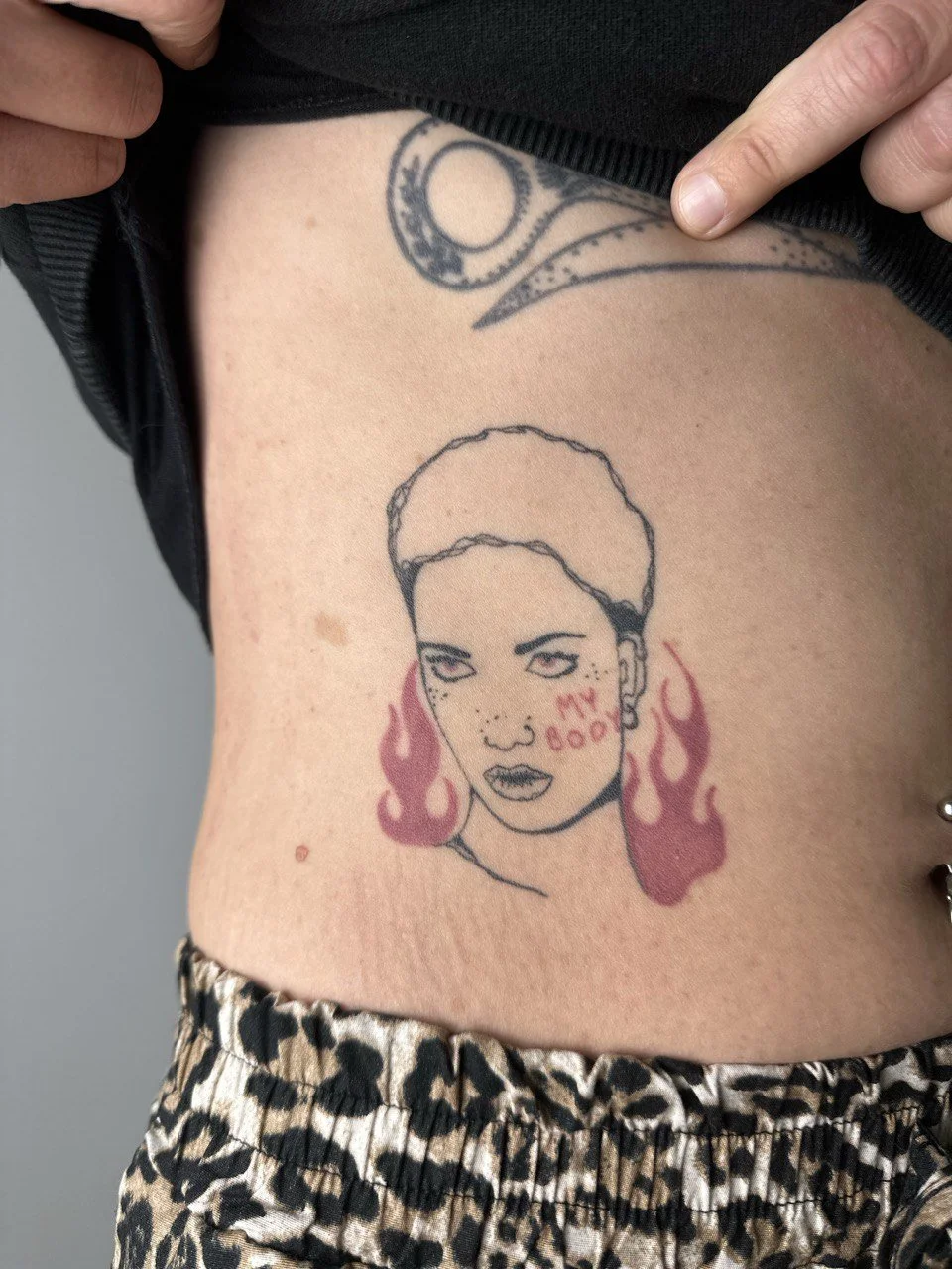 Tattoo eines Gesichts einer Frau mit kurzen Haaren, Flammen und dem Text "My Body" auf der rechten Wange.
