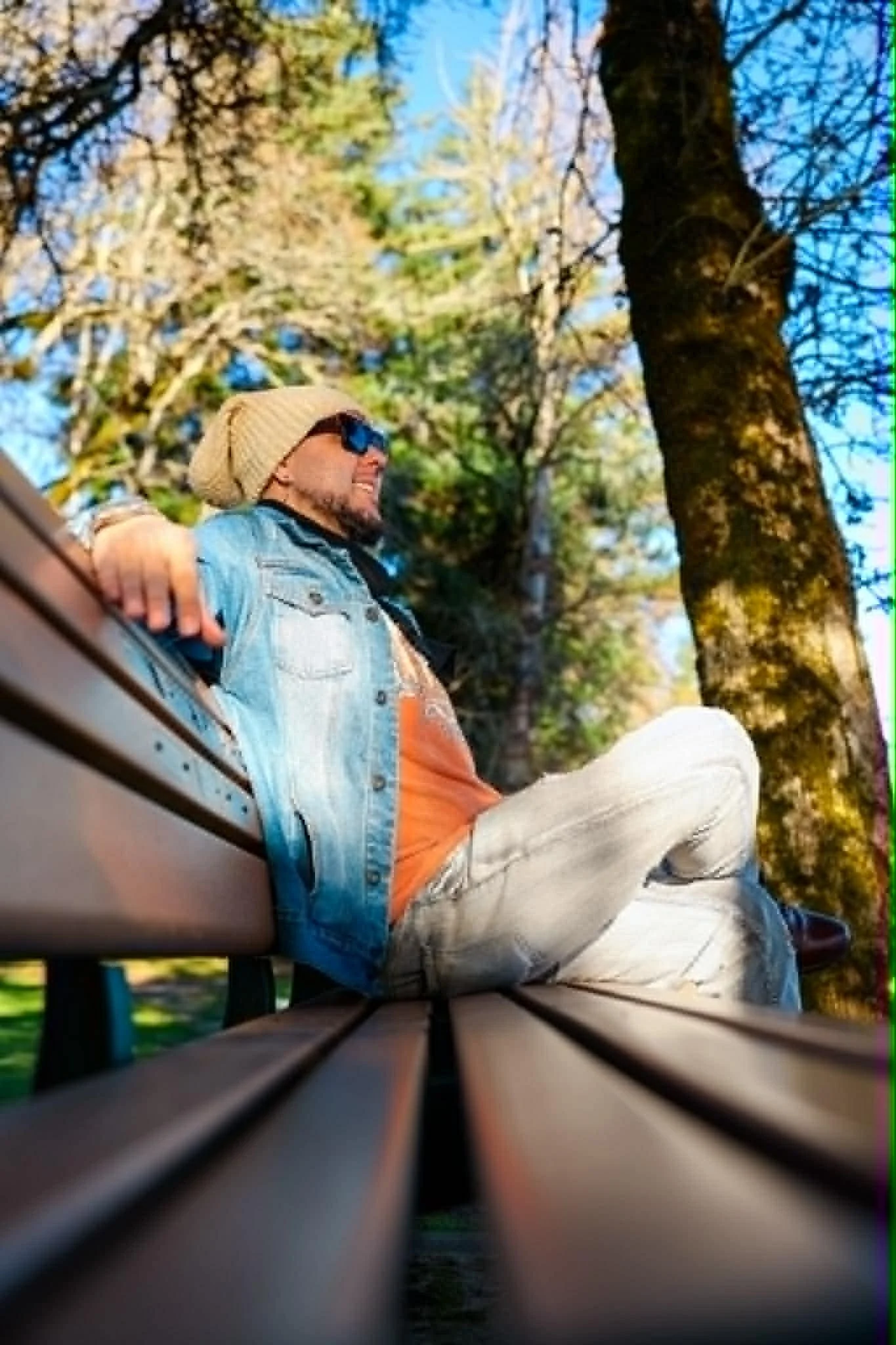 A man wearing sunglasses, a beige beanie, a light denim jacket, and light-colored pants sitting on a park bench surrounded by trees with green and brown leaves during autumn.