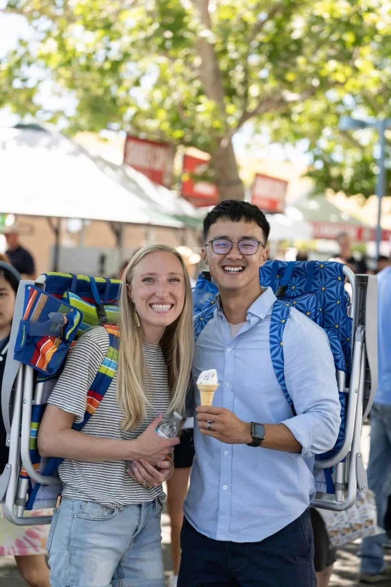 A smiling woman and man carrying large backpacks at an outdoor event, with trees and tents in the background. The man is holding a vanilla ice cream cone.