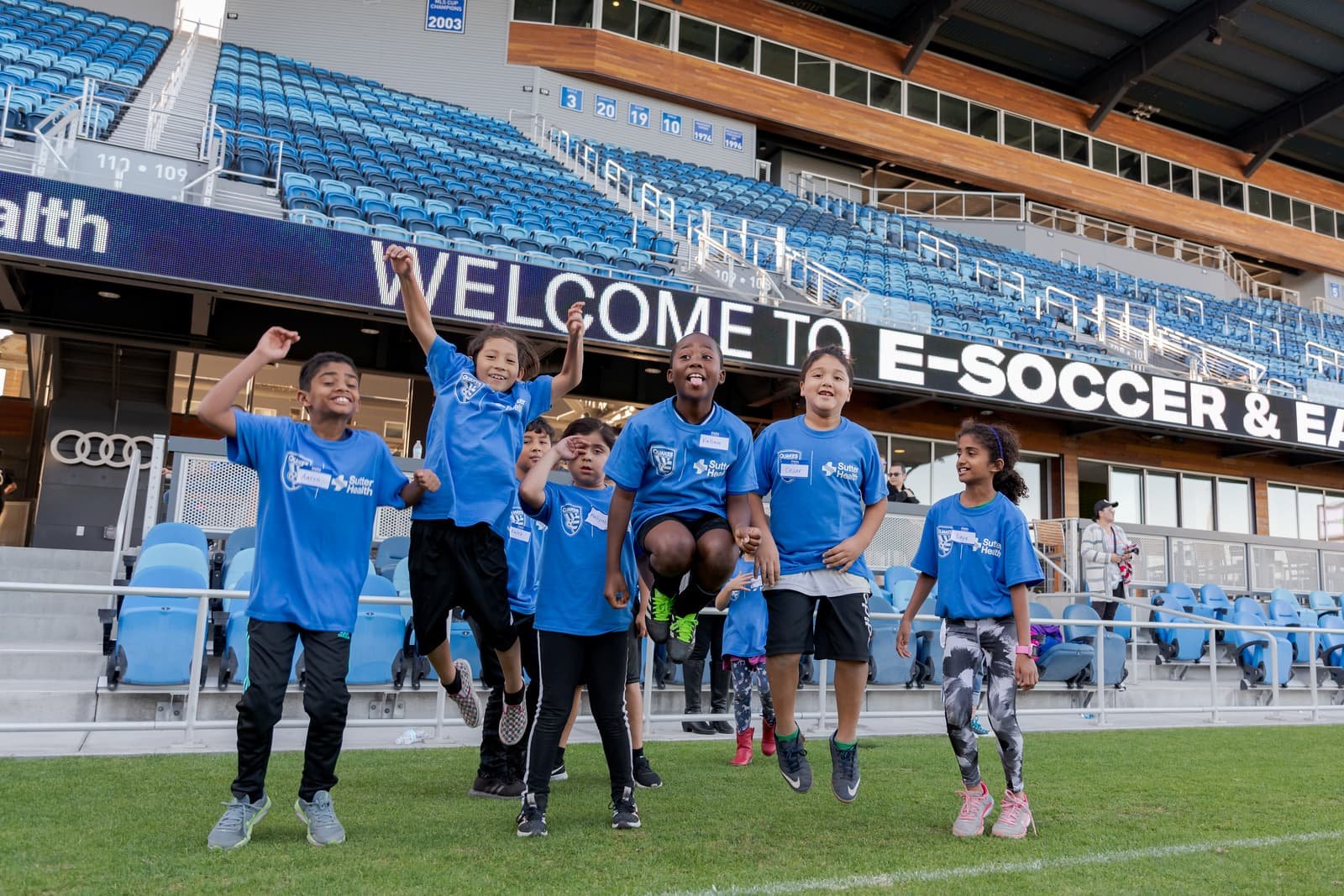 Children in blue sports shirts playing and jumping on a soccer field in a stadium, with blue seats and a large digital sign that reads 'WELCOME TO E- SOCCER & E' in the background.
