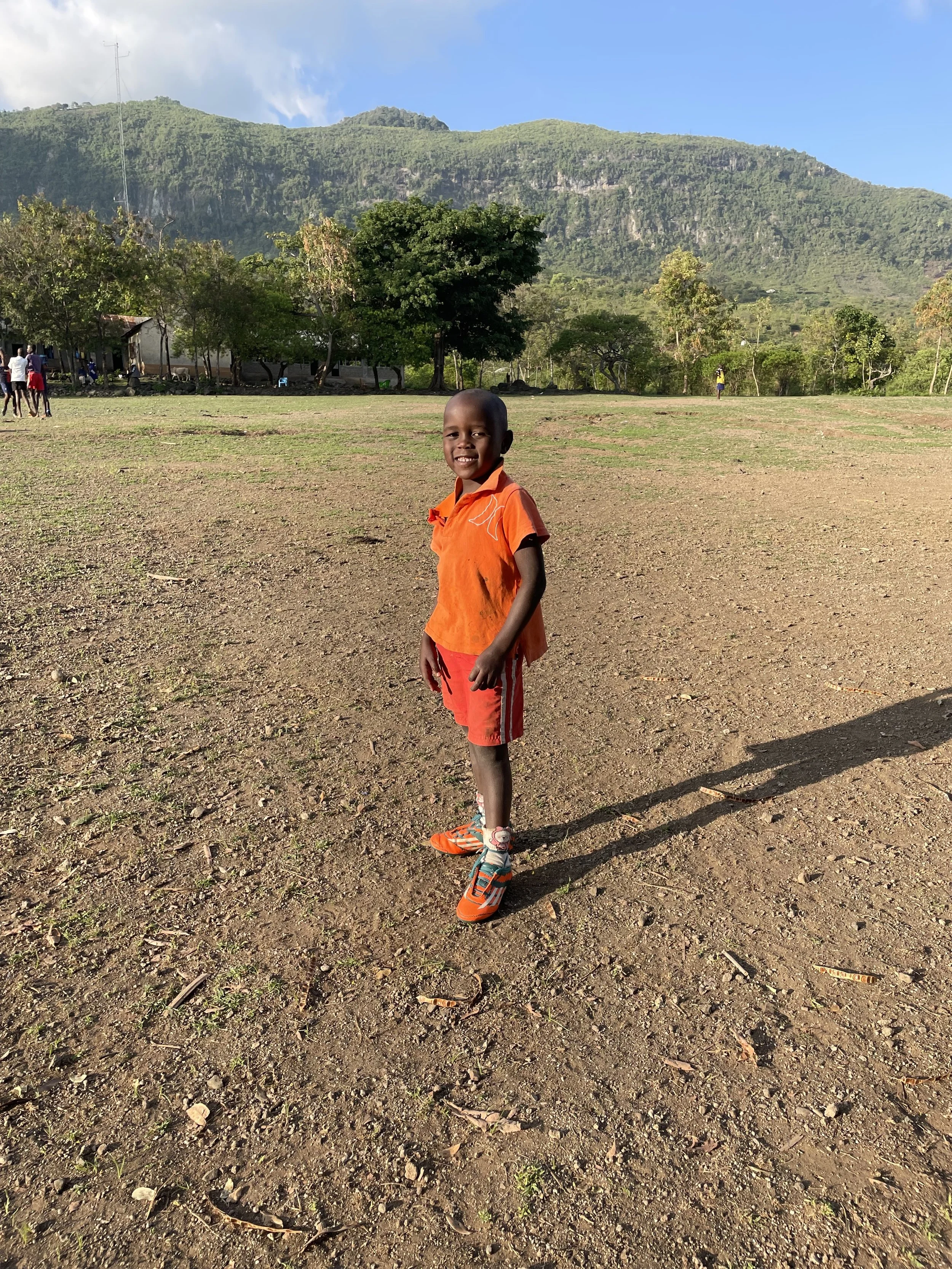 A young boy smiling, standing on a dirt field with mountains and trees in the background, dressed in an orange shirt, red shorts, and orange sneakers.
