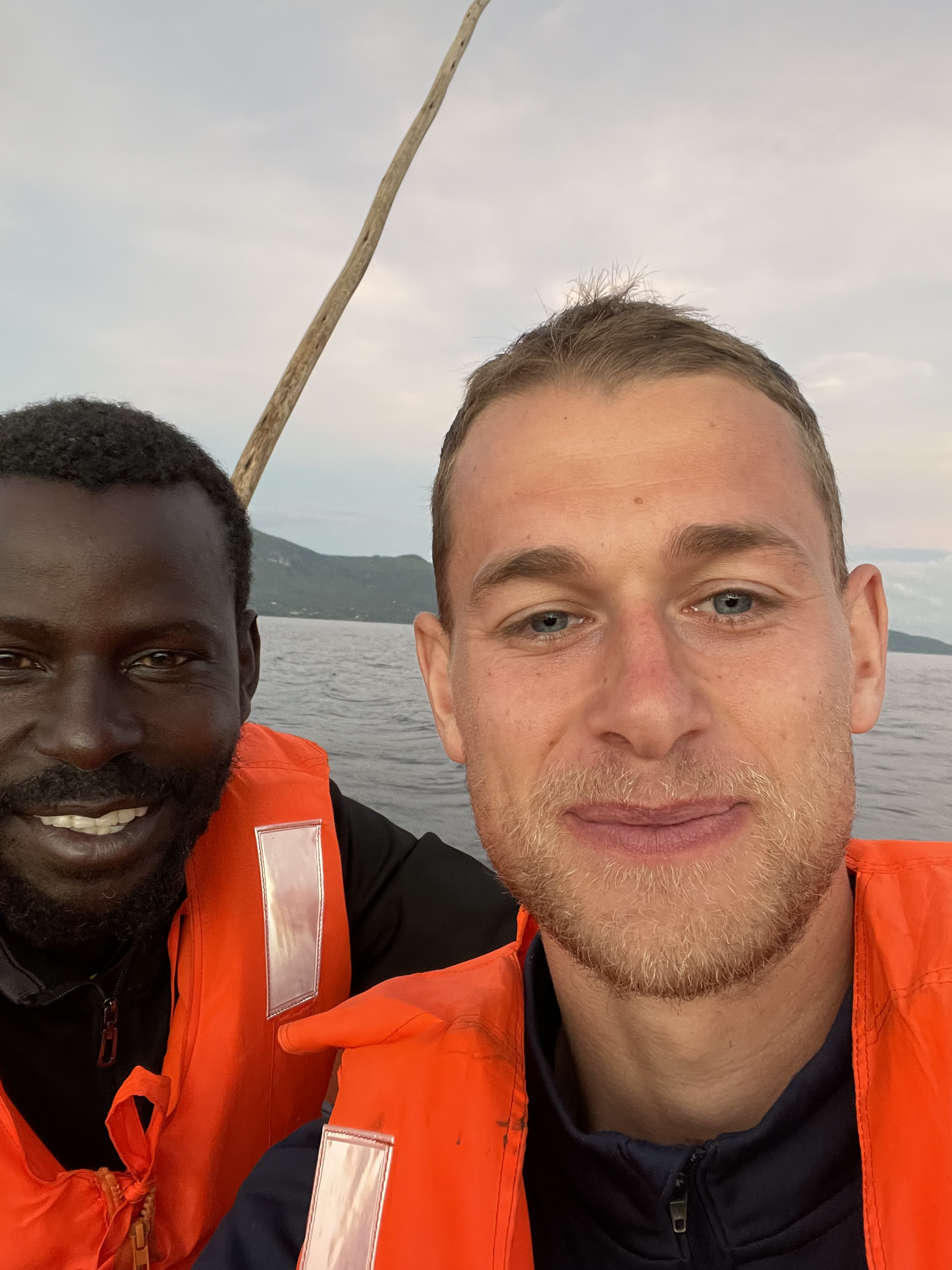 Two men wearing orange life jackets smiling for a selfie on a boat with water and mountains in the background.