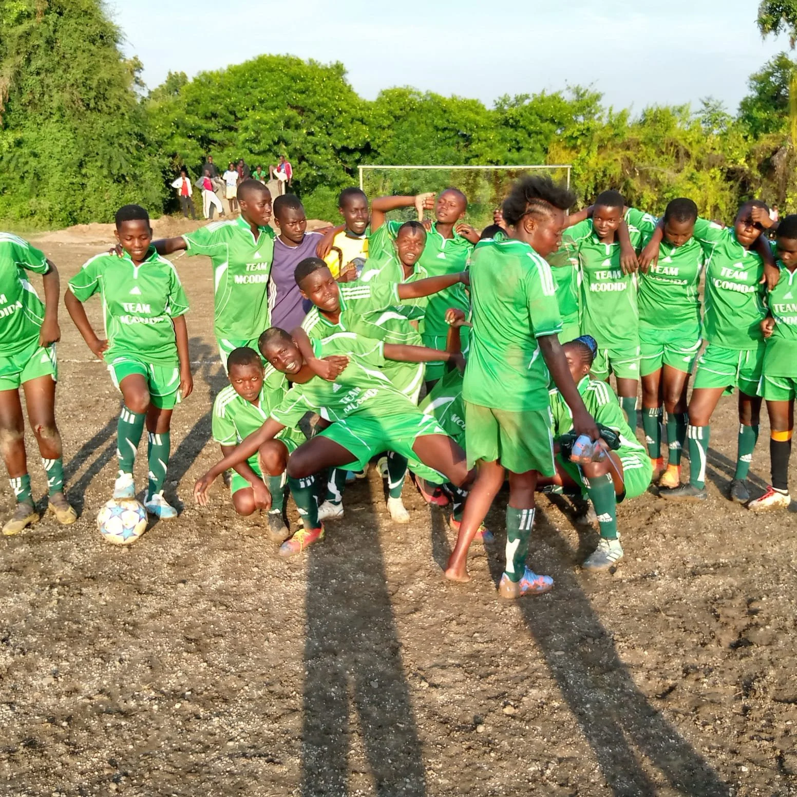 A group of young girls in green soccer uniforms standing on a dirt field, with trees and spectators in the background.