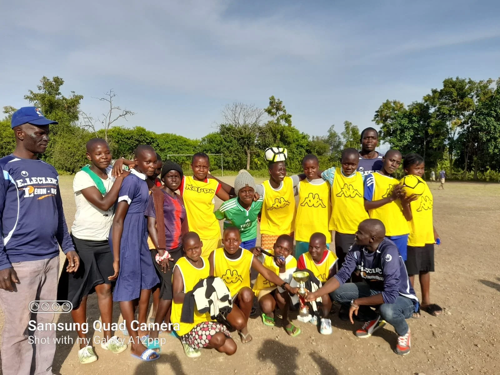 Group of children and coaches on a soccer field, some children wearing yellow jerseys, with a trophy and soccer balls, after a match or tournament, outdoors on a sunny day.