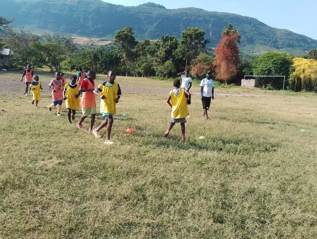 Children playing soccer on a grassy field with mountains and trees in the background, some children wearing yellow pinnies, some adults supervising.