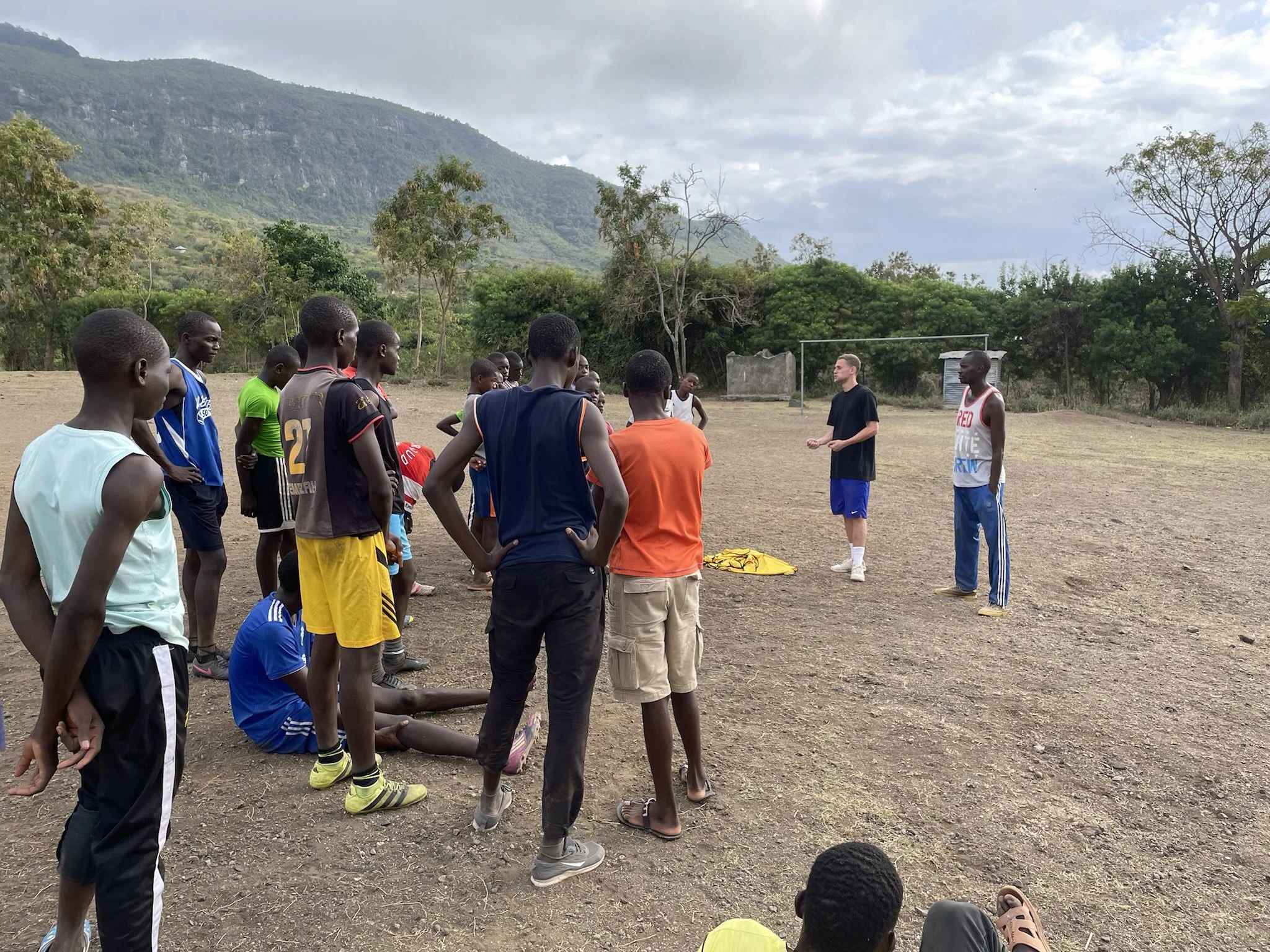 A group of young men in sportswear gathered on a dirt field, listening to a coach giving instructions, with a backdrop of green hills and trees.