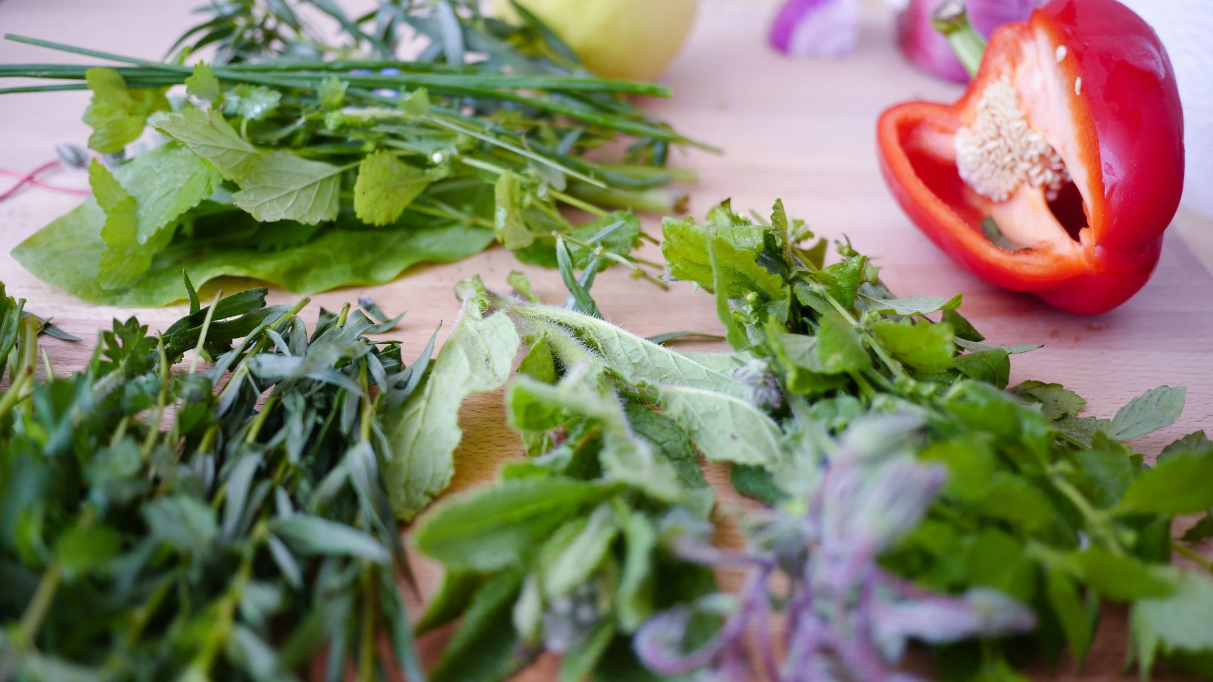 Fresh herbs and vegetables on a wooden surface, including red bell pepper sliced in half, and various green herbs.