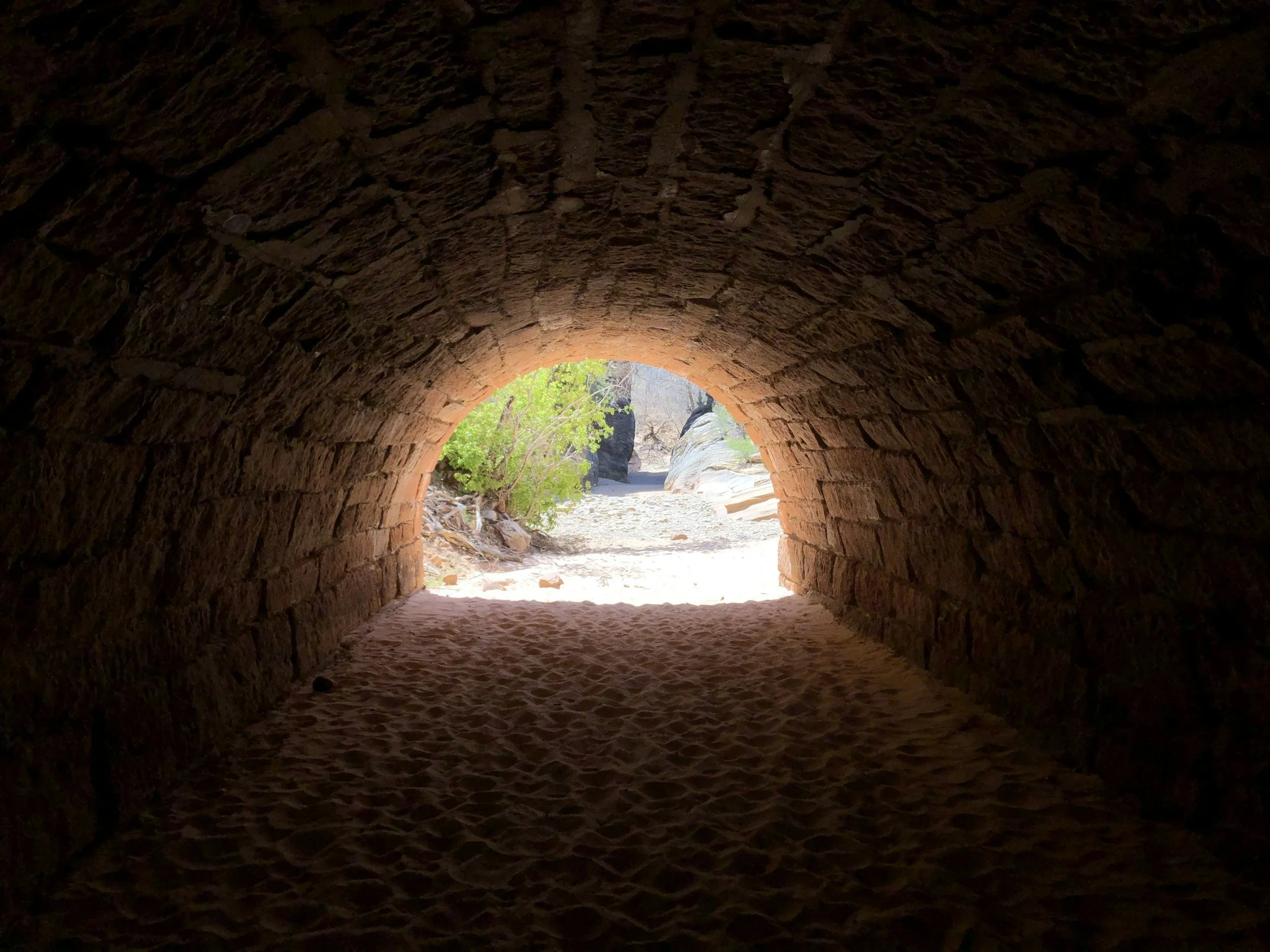 View through a tunnel made of stone bricks, with bright sunlight and green bushes outside.