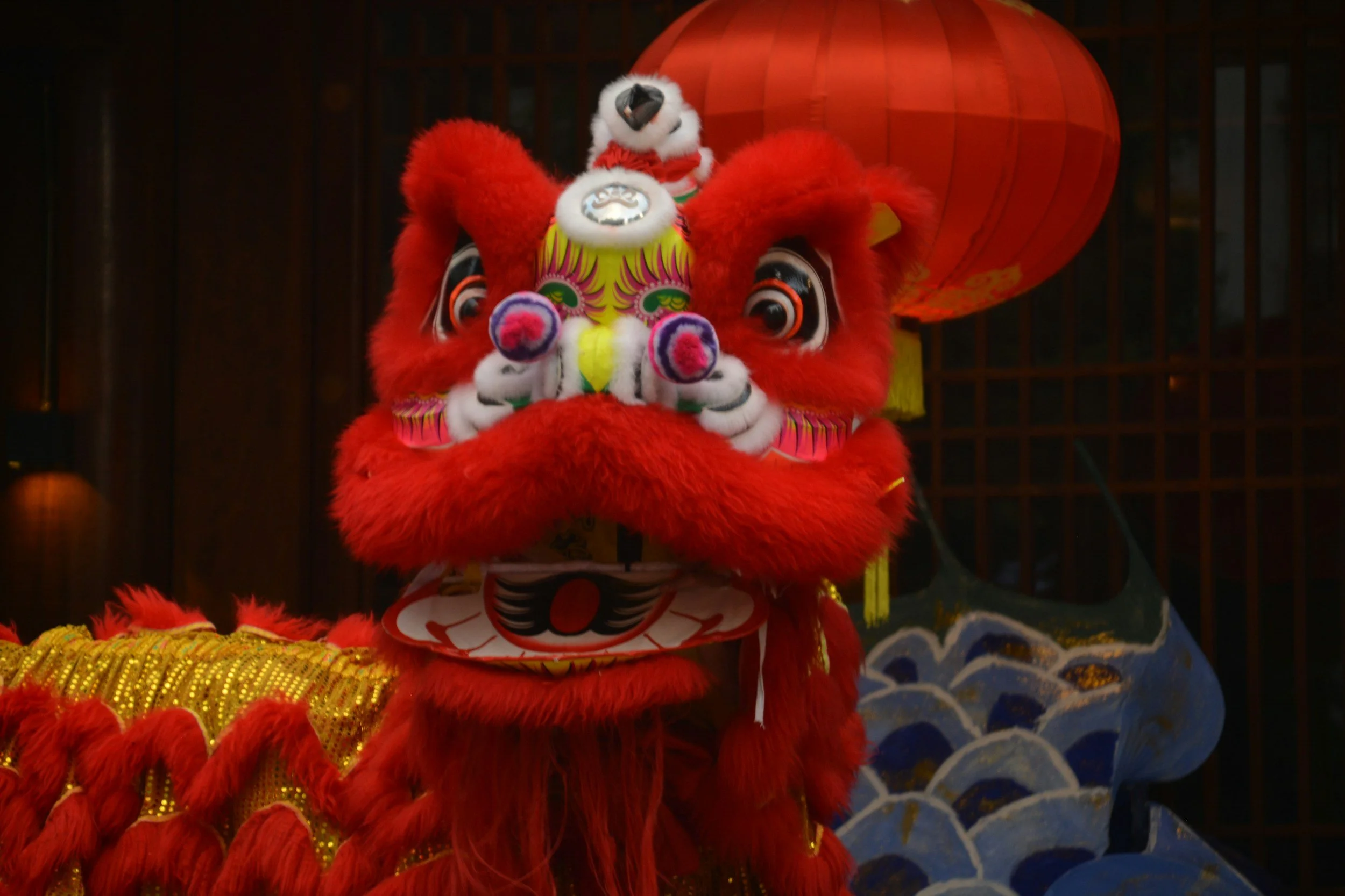 Red lion dance costume with a large, detailed head and big eyes, adorned with traditional decorations, with a red lantern and painted waves in the background.