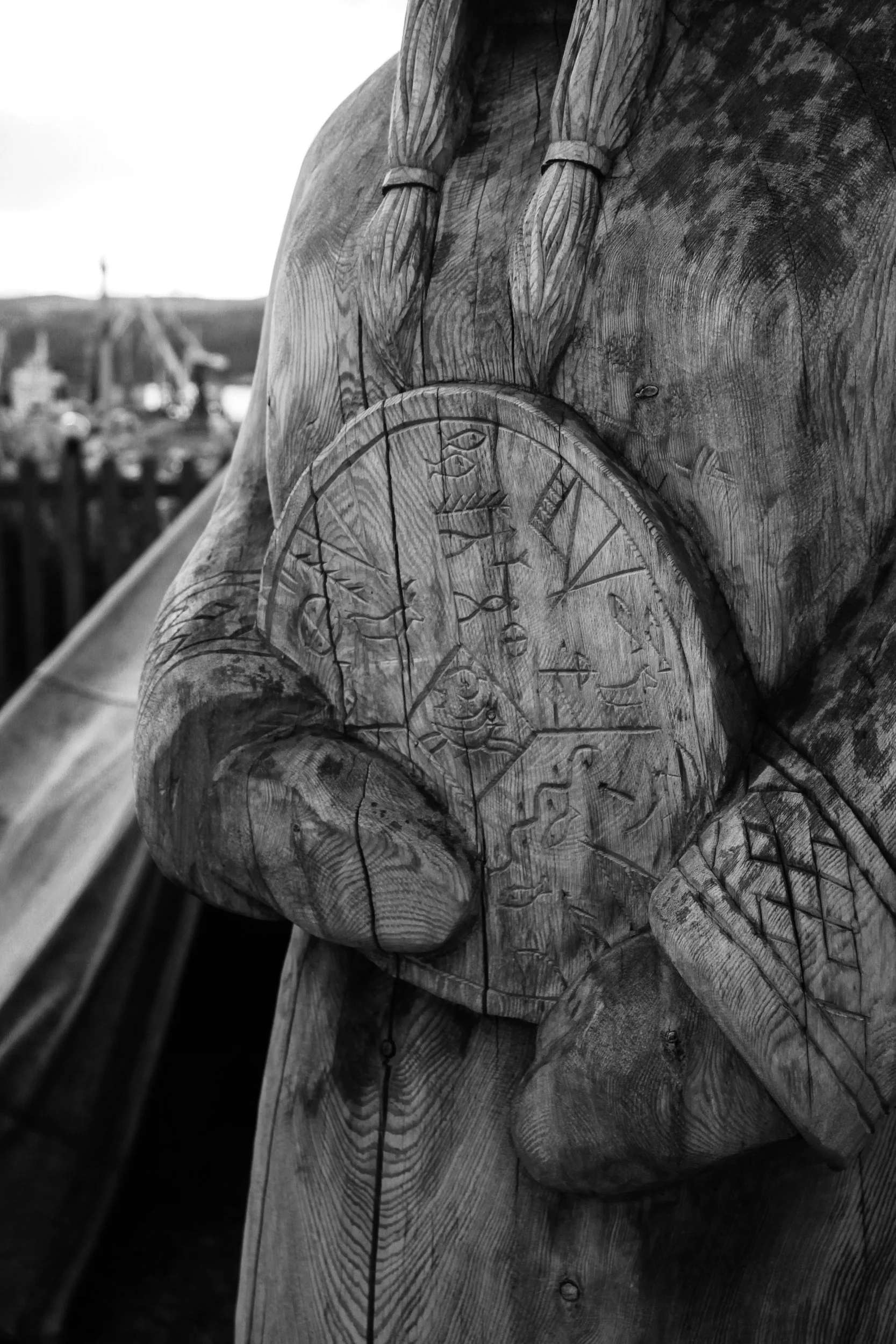 Close-up of a wooden sculpture of a hand holding a clock, carved with detailed symbols and Roman numerals. In the background, a blurred outdoor setting with structures and people.