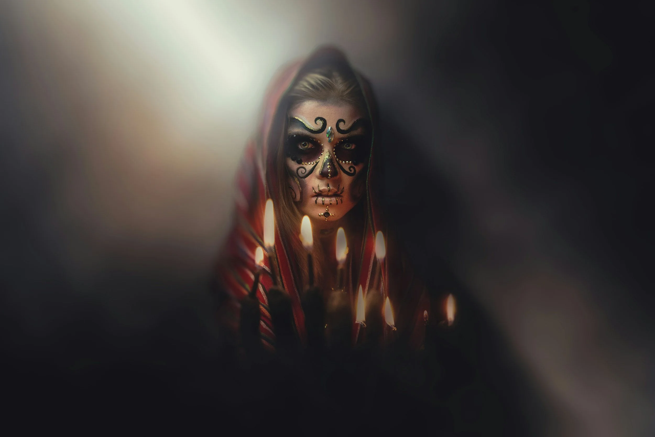 A person with sugar skull makeup and face paint, wearing a hooded red and black garment, surrounded by lit candles in a dark setting.