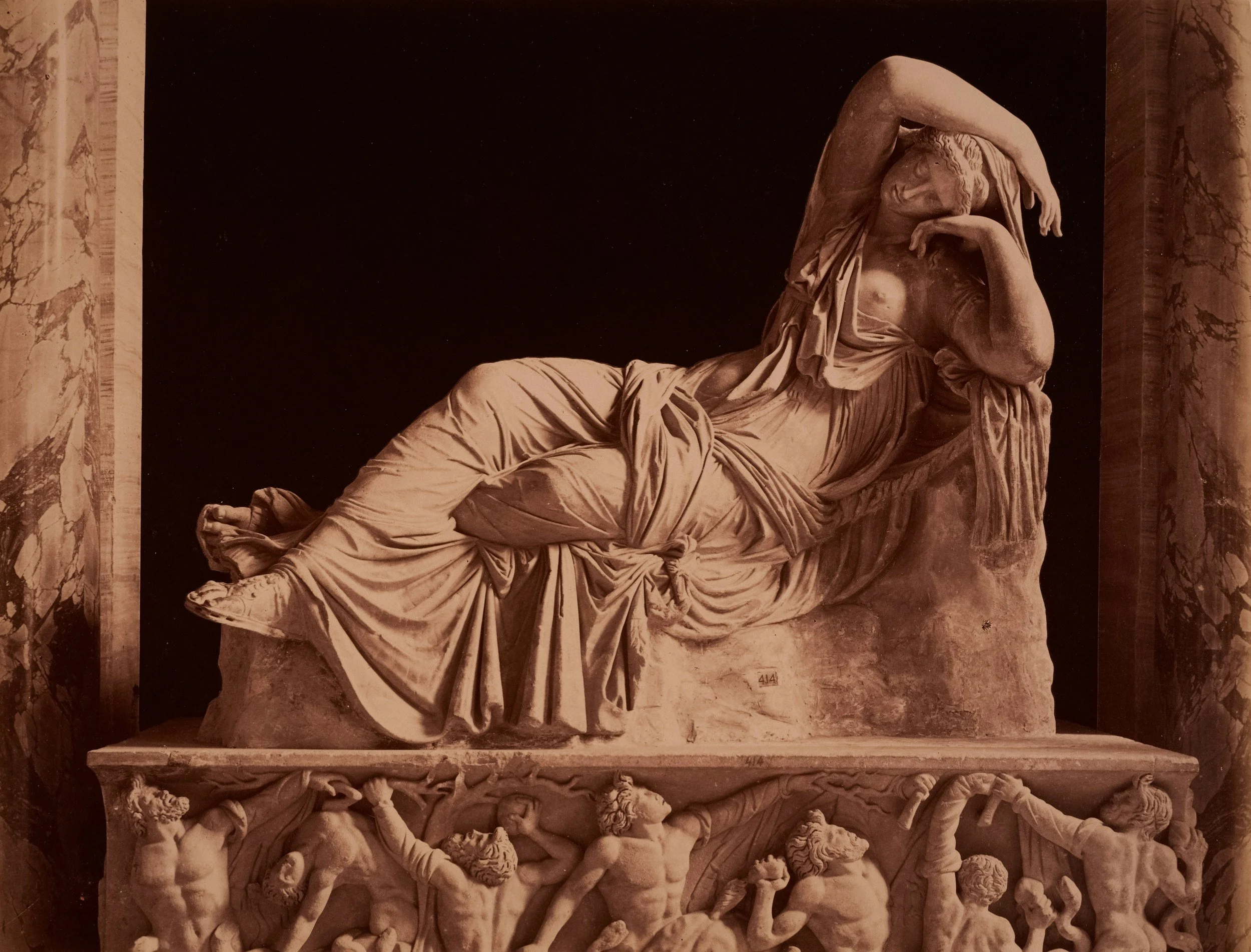 A classical marble sculpture of a reclining woman with sheaves of fabric, resting her head on her left arm, on a pedestal with carved figures below.