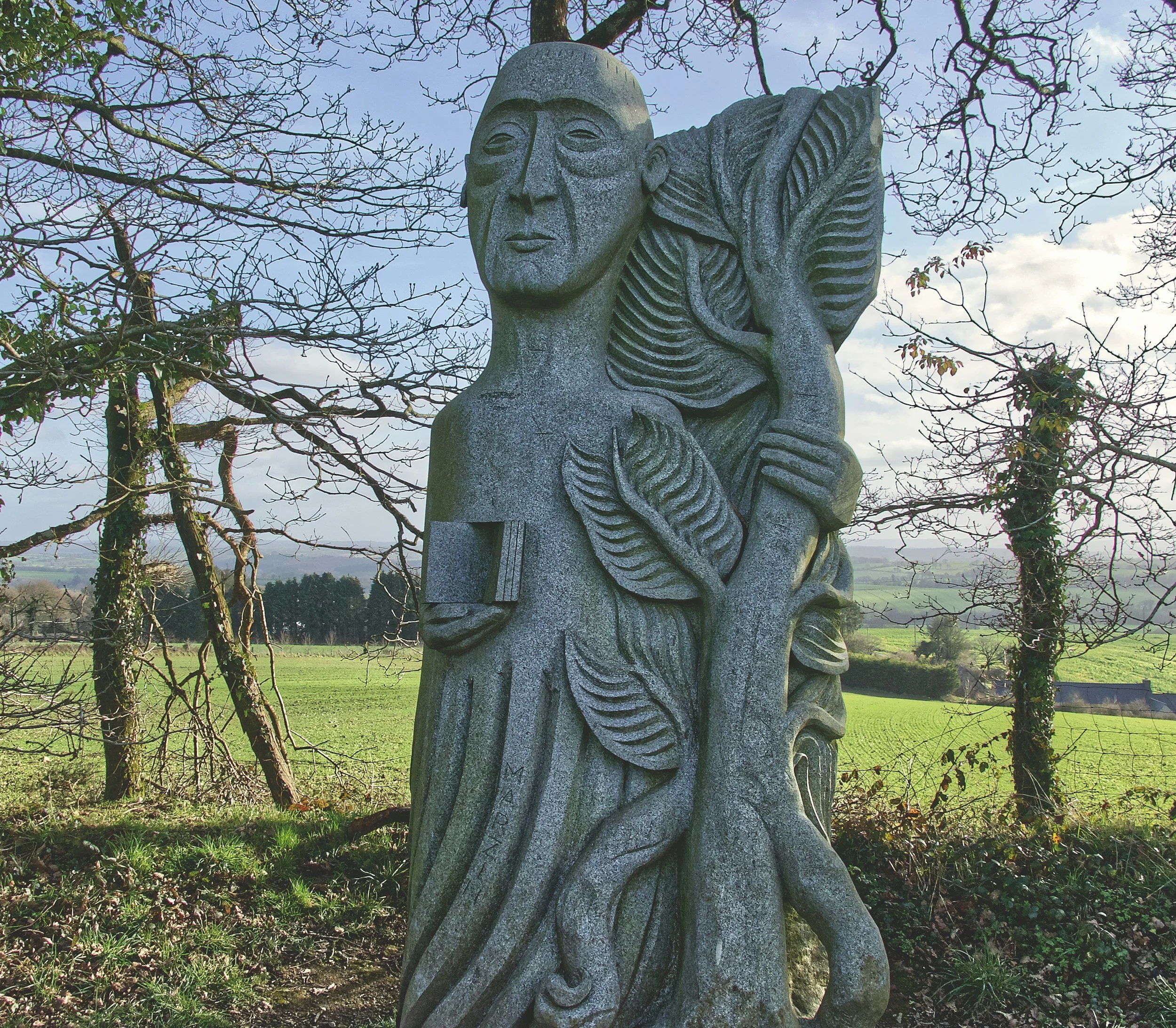 A stone sculpture of a contemplative man with a book in his pocket, surrounded by carved leaves and vines, set outdoors with leafless trees and a green field in the background under a cloudy sky.