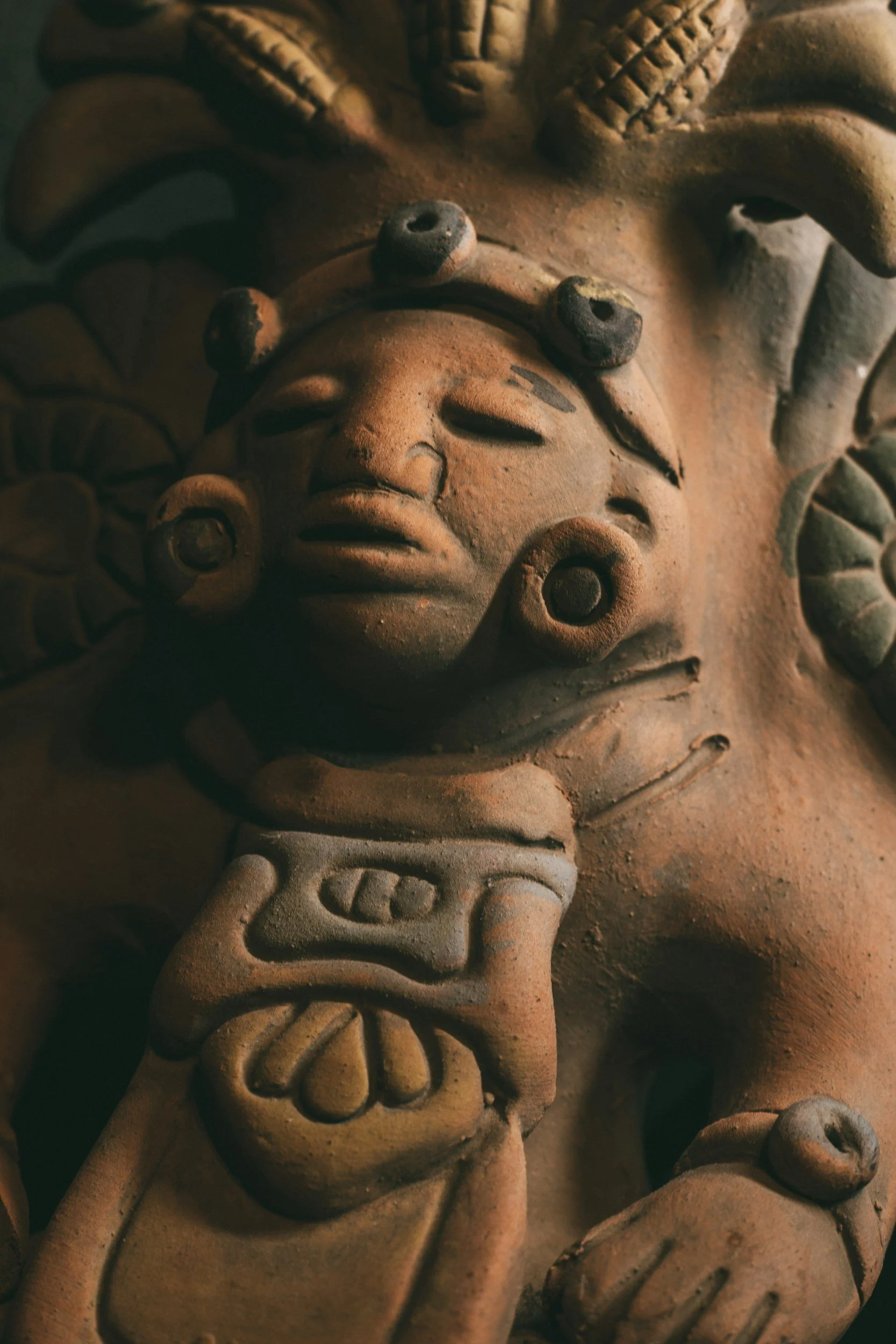 Close-up of a terracotta sculpture of a human face surrounded by decorative elements, including leaves and geometric patterns.