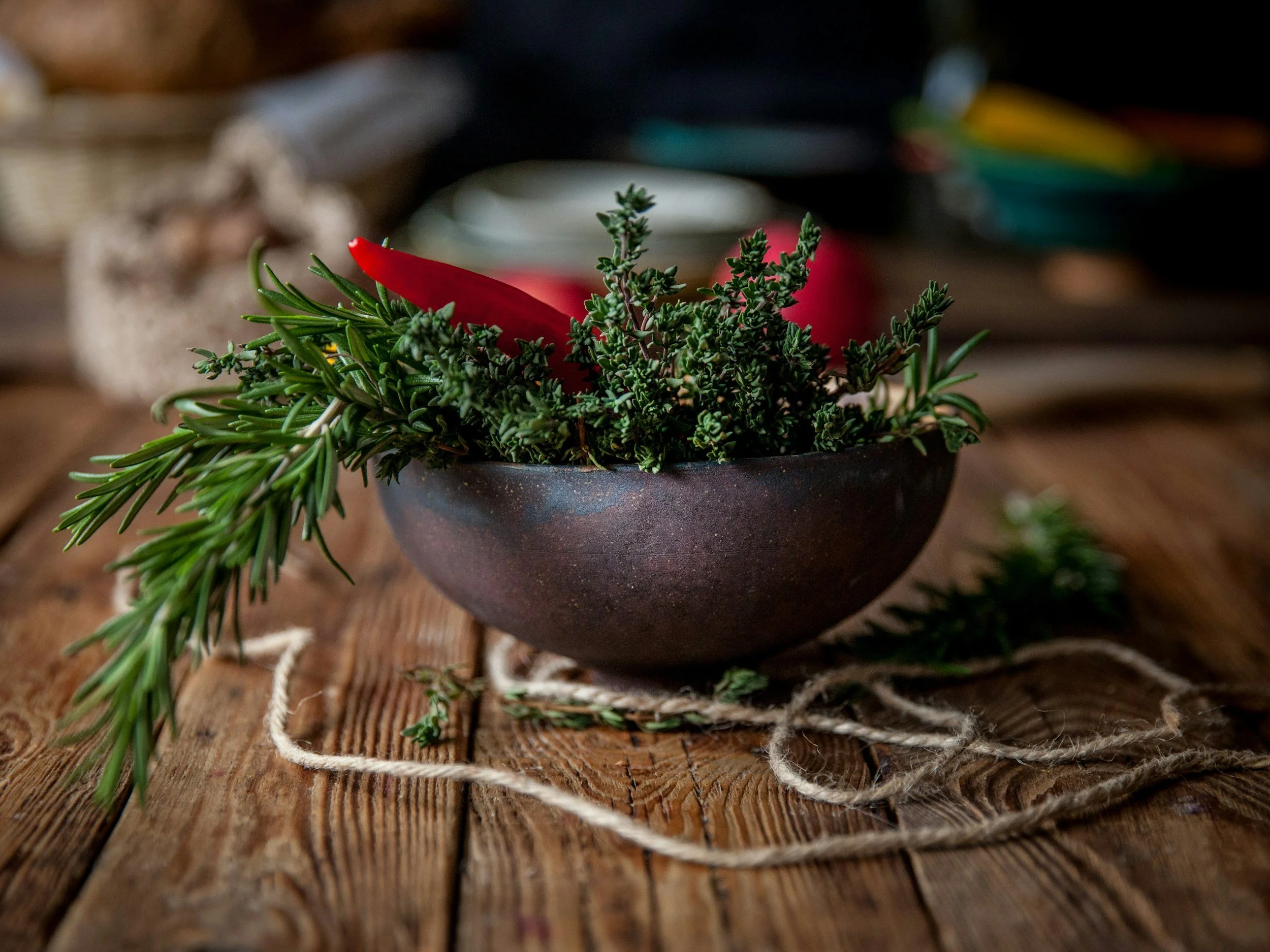 A black ceramic bowl containing green sprigs of herbs and a red chili pepper, placed on a wooden surface with twine around it.