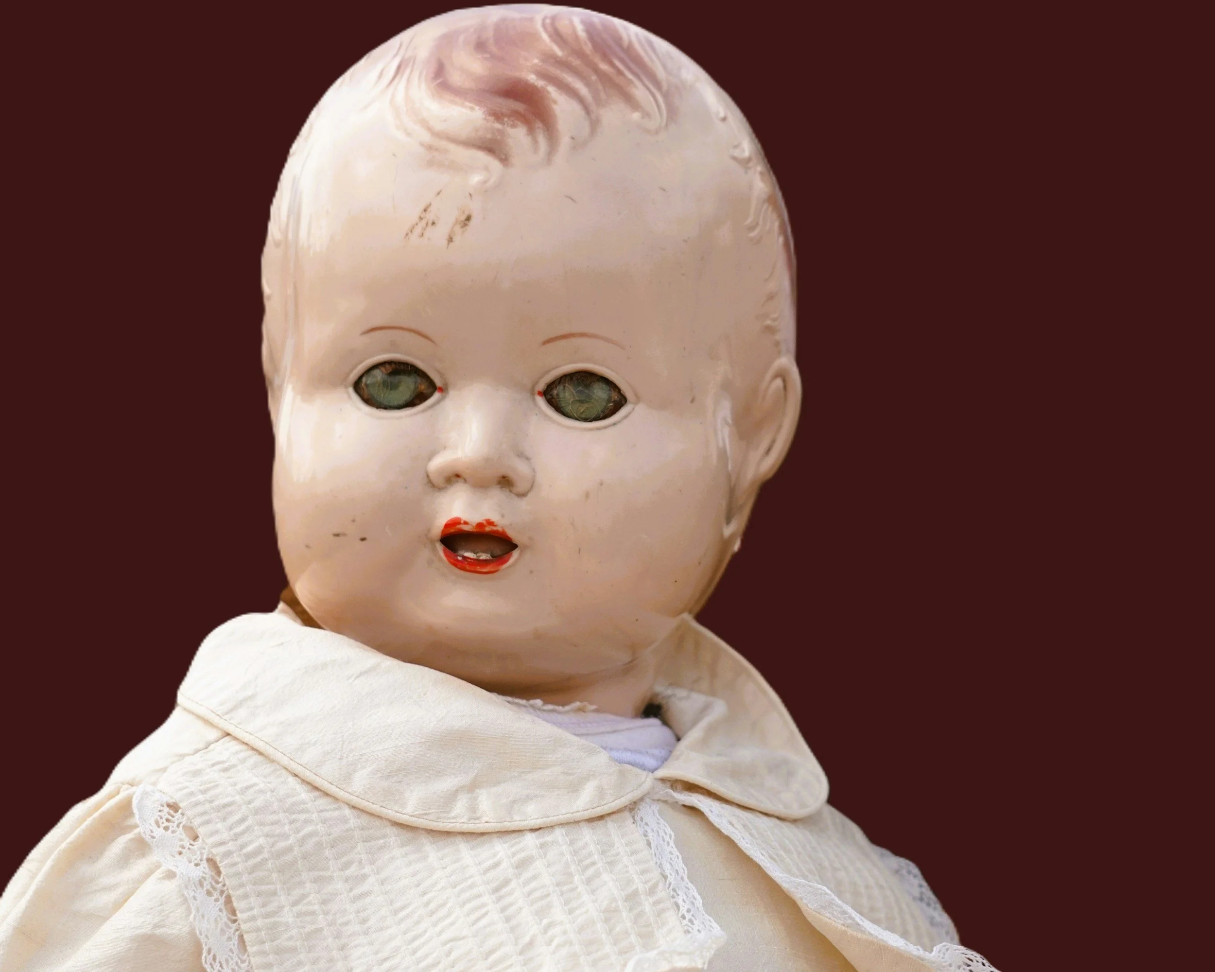 An antique doll with a cracked porcelain face, missing eyes, and a chipped mouth, wearing a white dress with lace details.