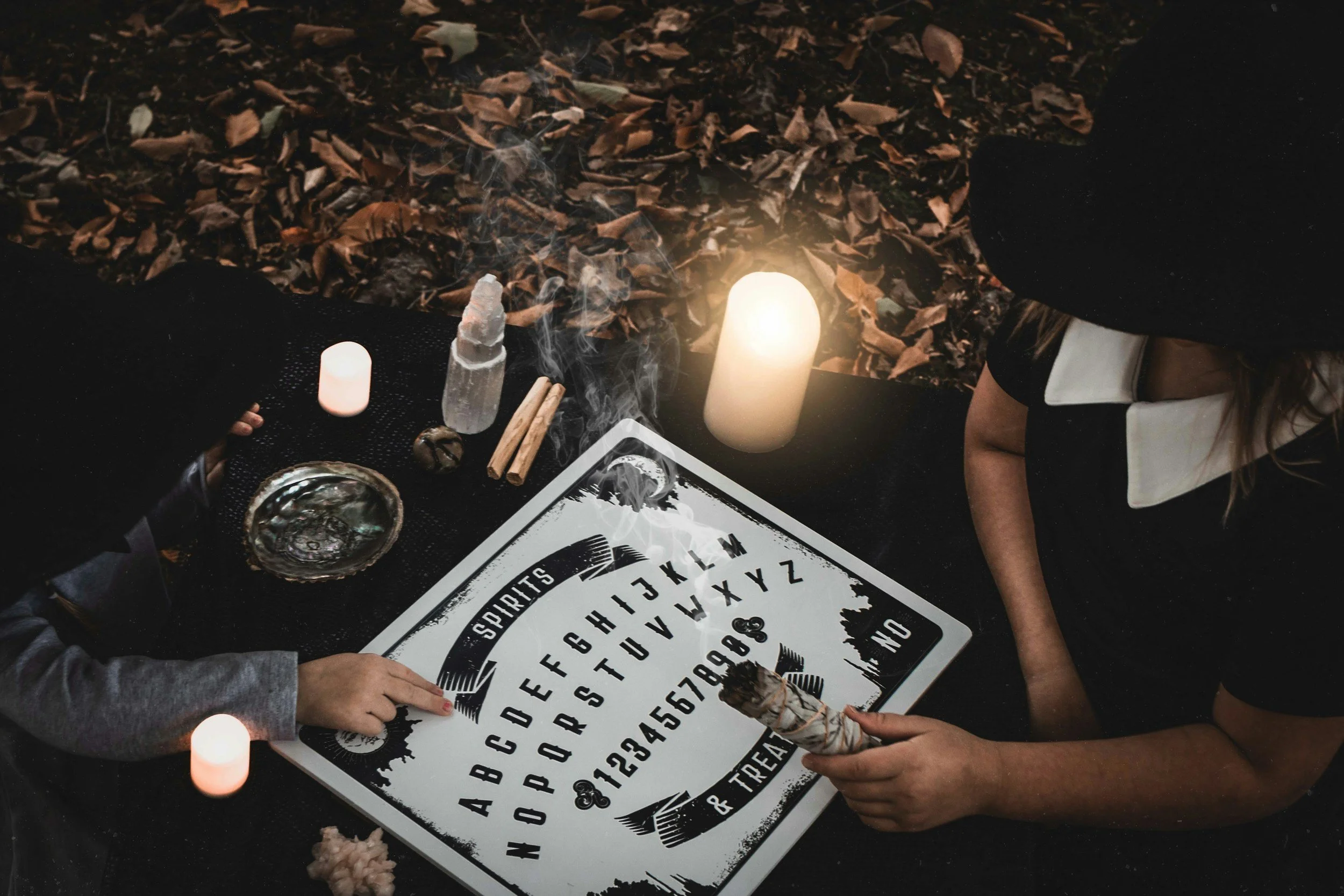 A person and a child are participating in a Ouija board session outdoors at night, surrounded by candles, crystals, and autumn leaves.