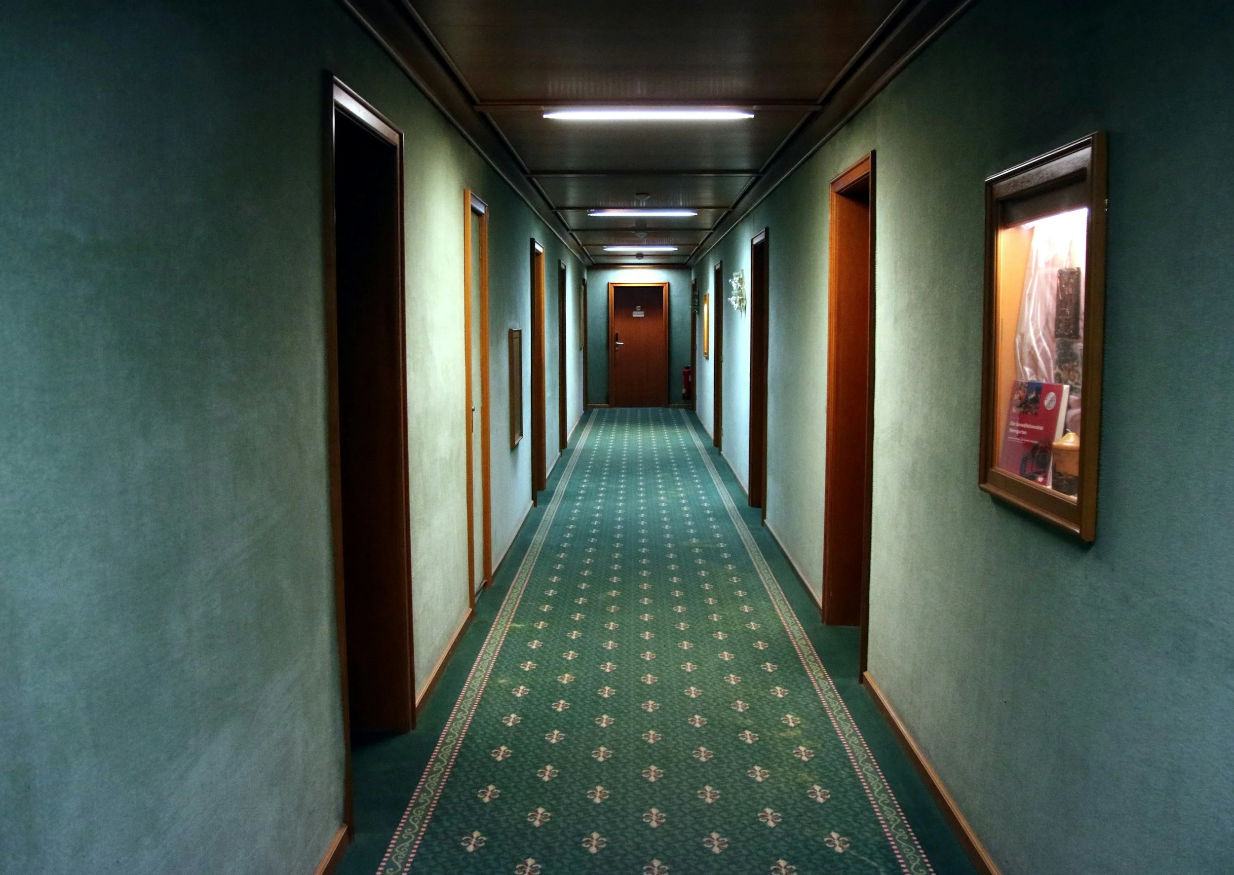 Empty hotel hallway with green walls, wooden door frames, and a patterned green carpet. Fluorescent ceiling lights and framed pictures on the walls.