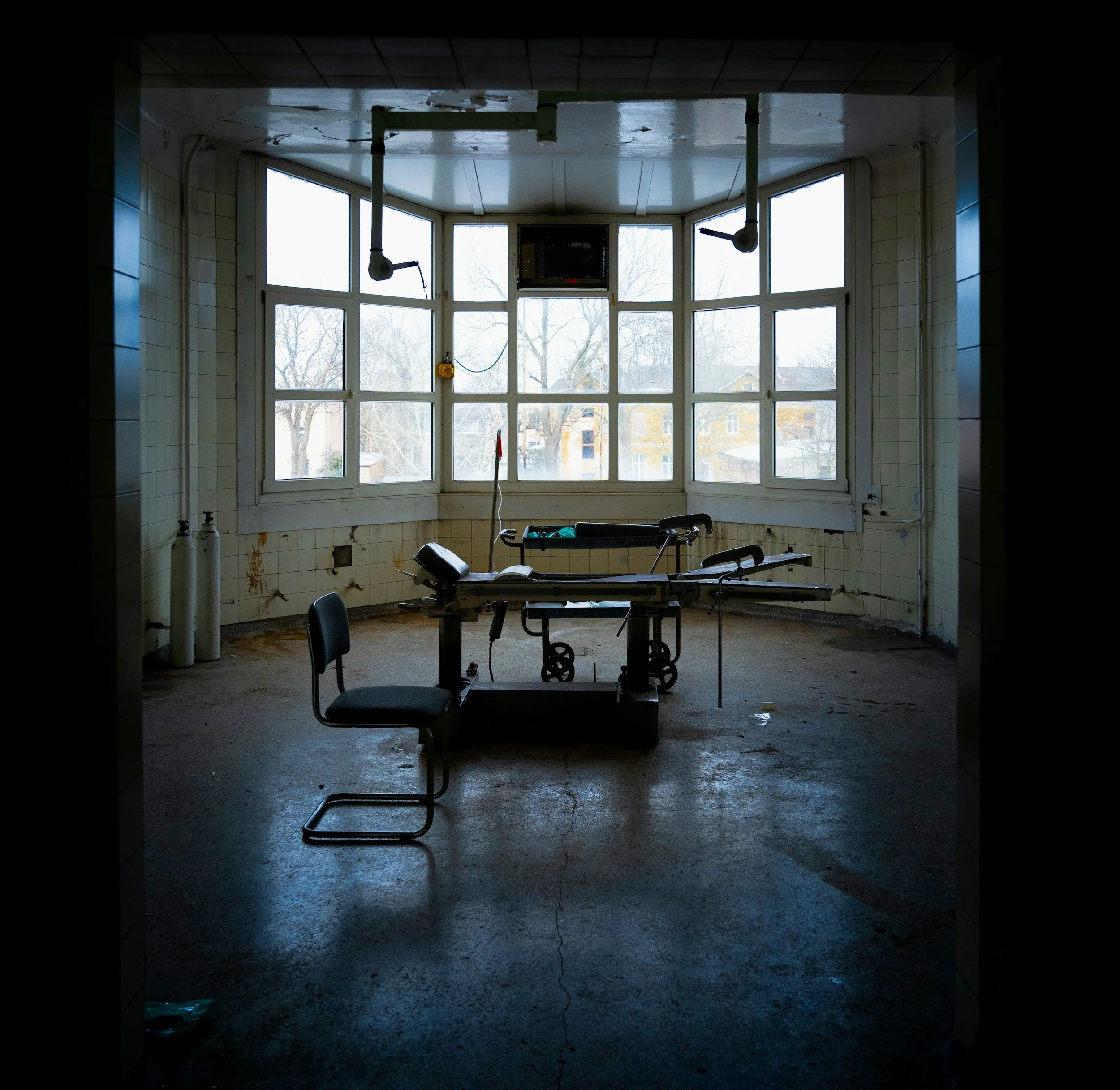 Empty hospital room with large windows, hospital bed, and a chair, appearing abandoned and in disrepair.