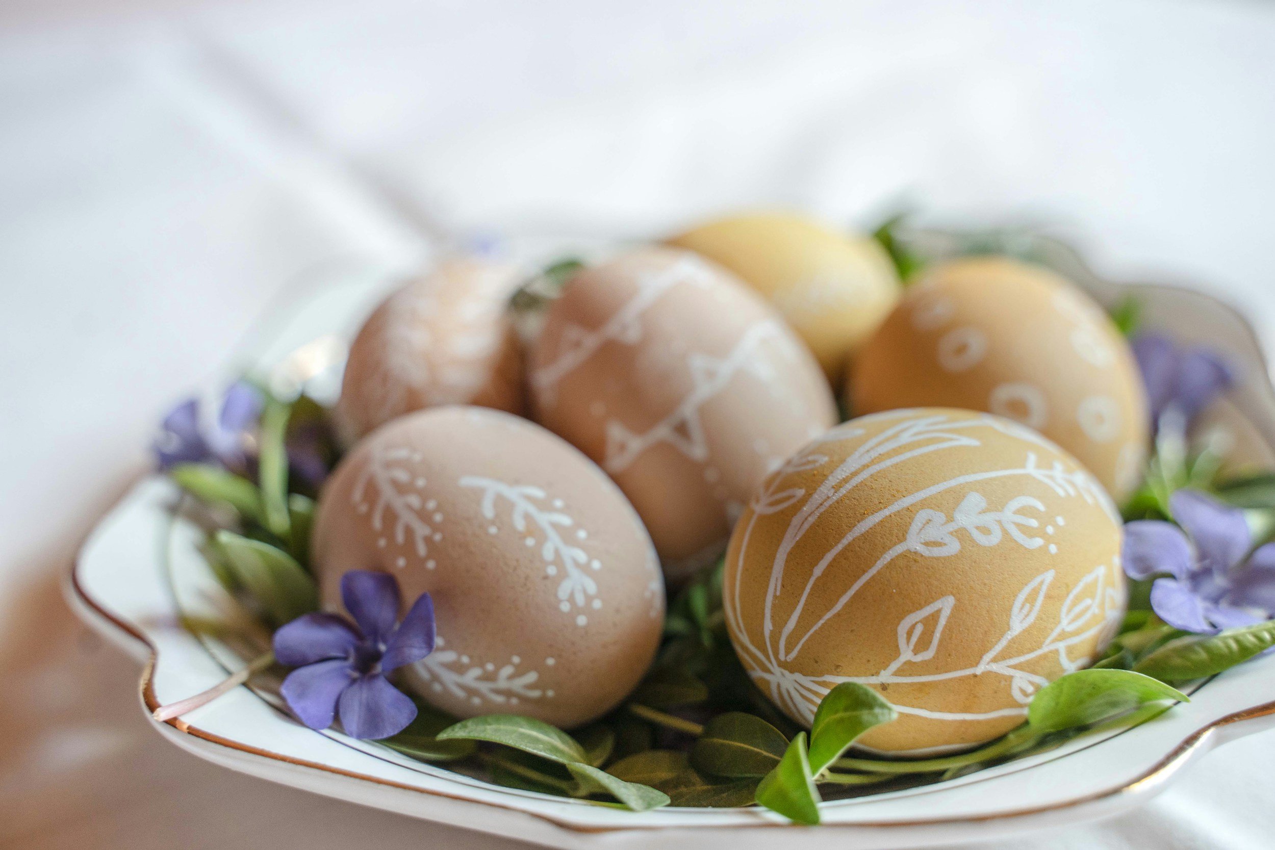 Decorative Easter eggs in pastel colors with white floral patterns, surrounded by green leaves and purple flowers on a white dish.