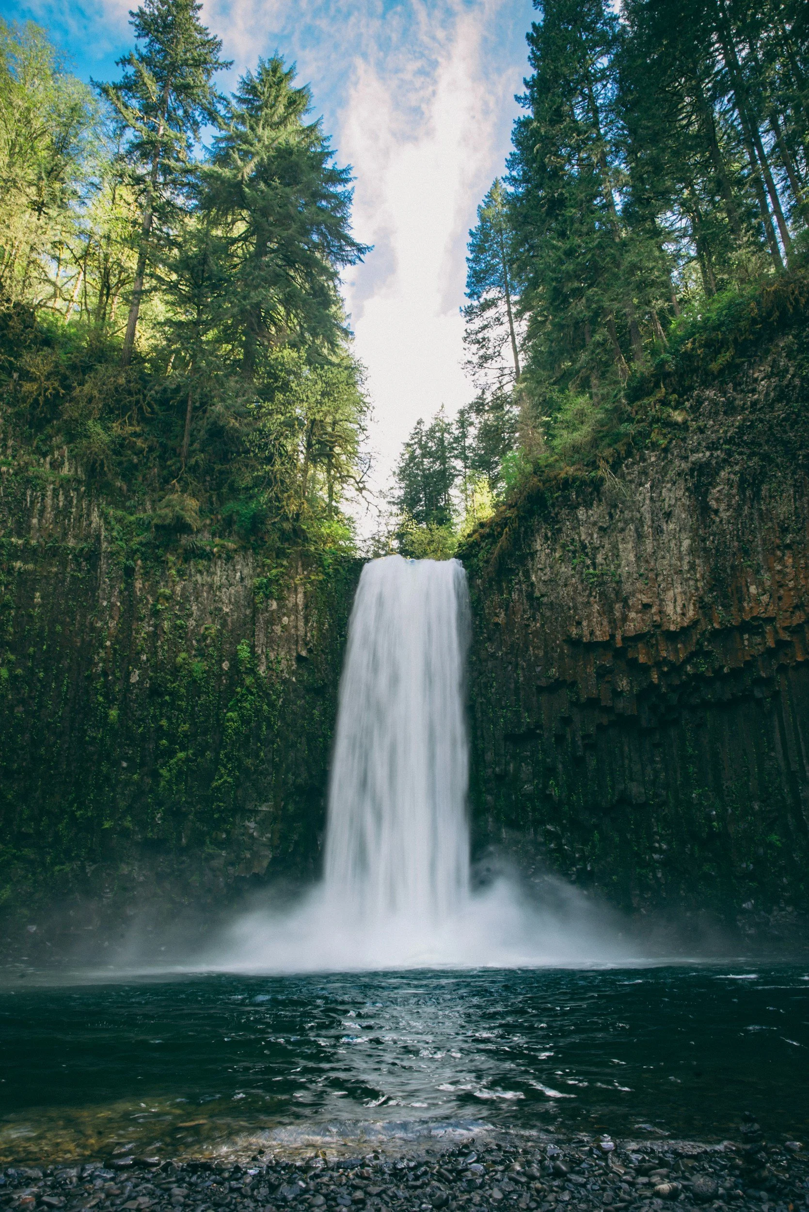 A waterfall cascading down a rocky cliff surrounded by tall evergreen trees under a partly cloudy sky.