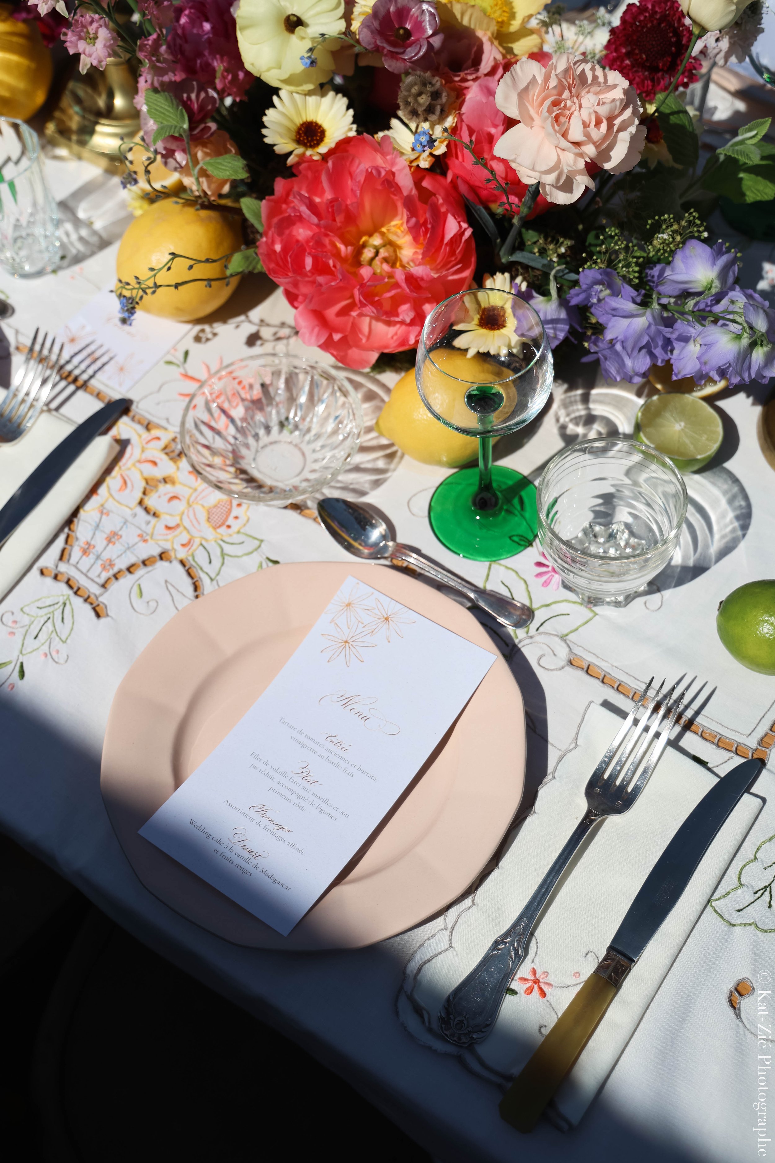 Table de réception élégante avec une nappe blanche, un menu, des couverts, un verre à vin avec une fleur dedans, un verre d'eau, des citrons, une citron vert, et un grand arrangement de fleurs colorées comprenant des roses, des pivoines, des lupins et des marguerites.