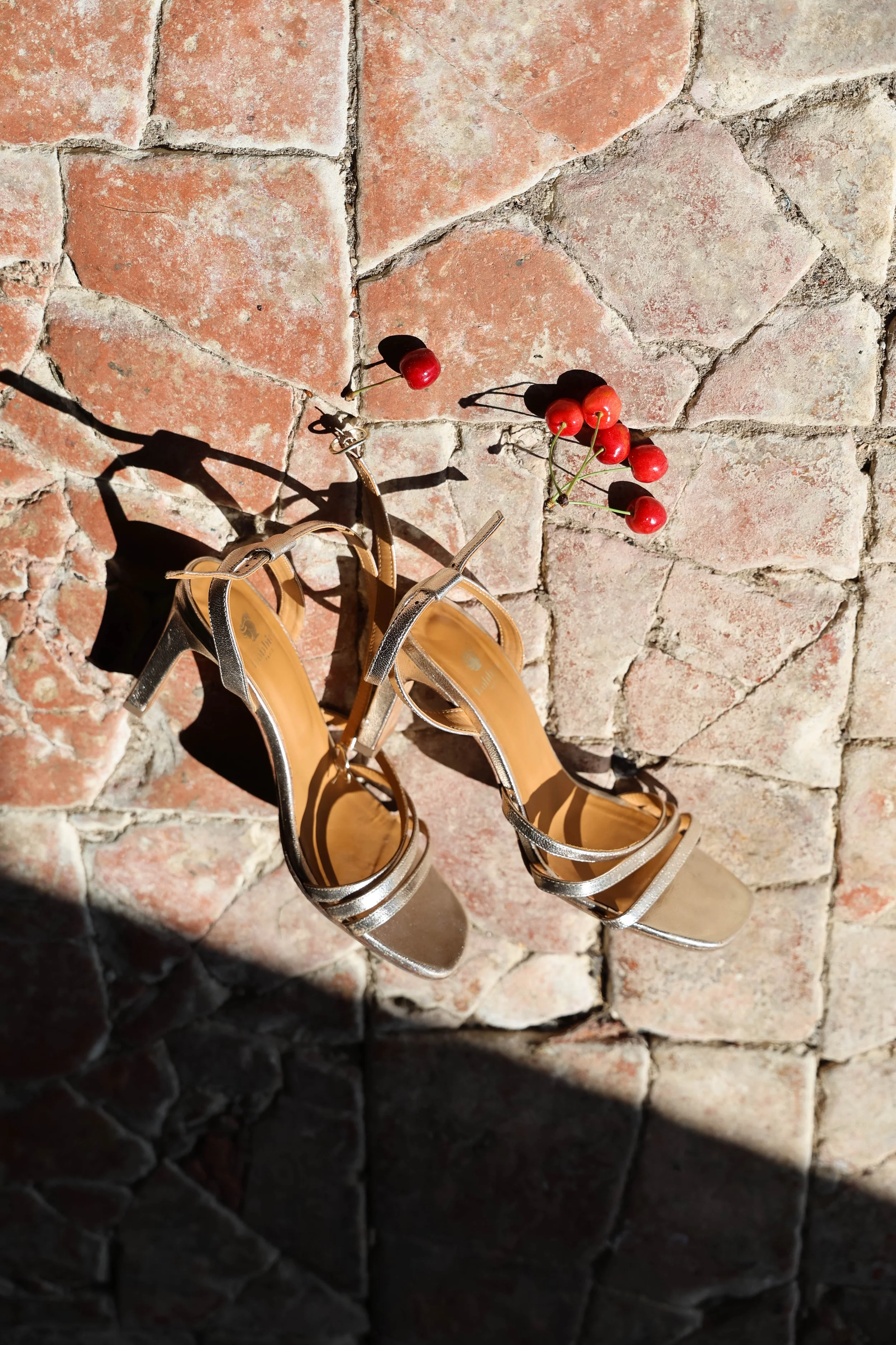 Une paire de chaussures à talons en coloris beige posée sur un sol en pavés en résine. Deux cerises rouges sont également posées à côté des chaussures, avec leurs ombres projetées sur le sol.