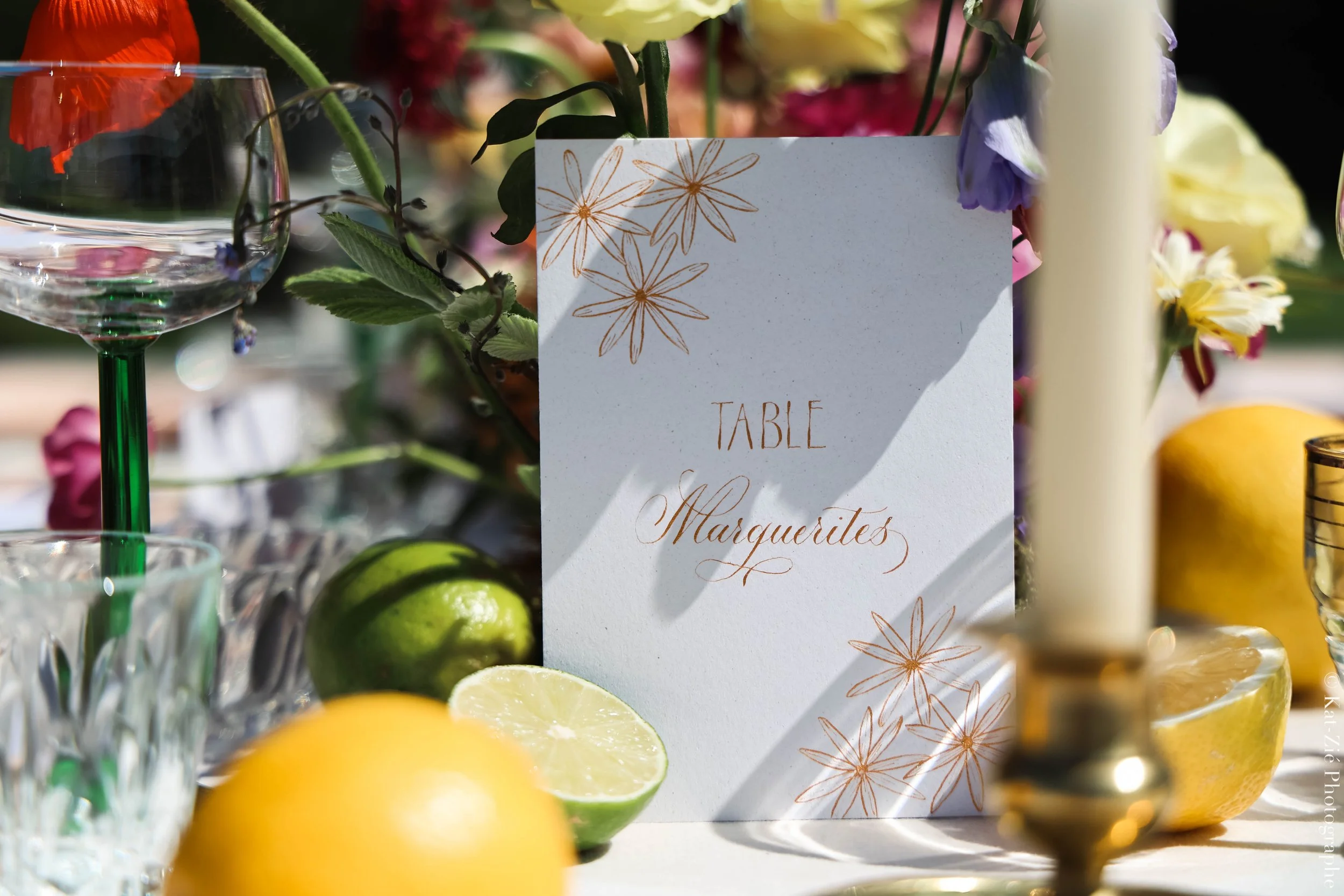 Une table décorée avec un menu sur fond blanc avec des fleurs dorées, des citrons coupés, un citron vert, un verre à vin avec une tige verte, des bougies, et des fleurs de différentes couleurs en arrière-plan.
