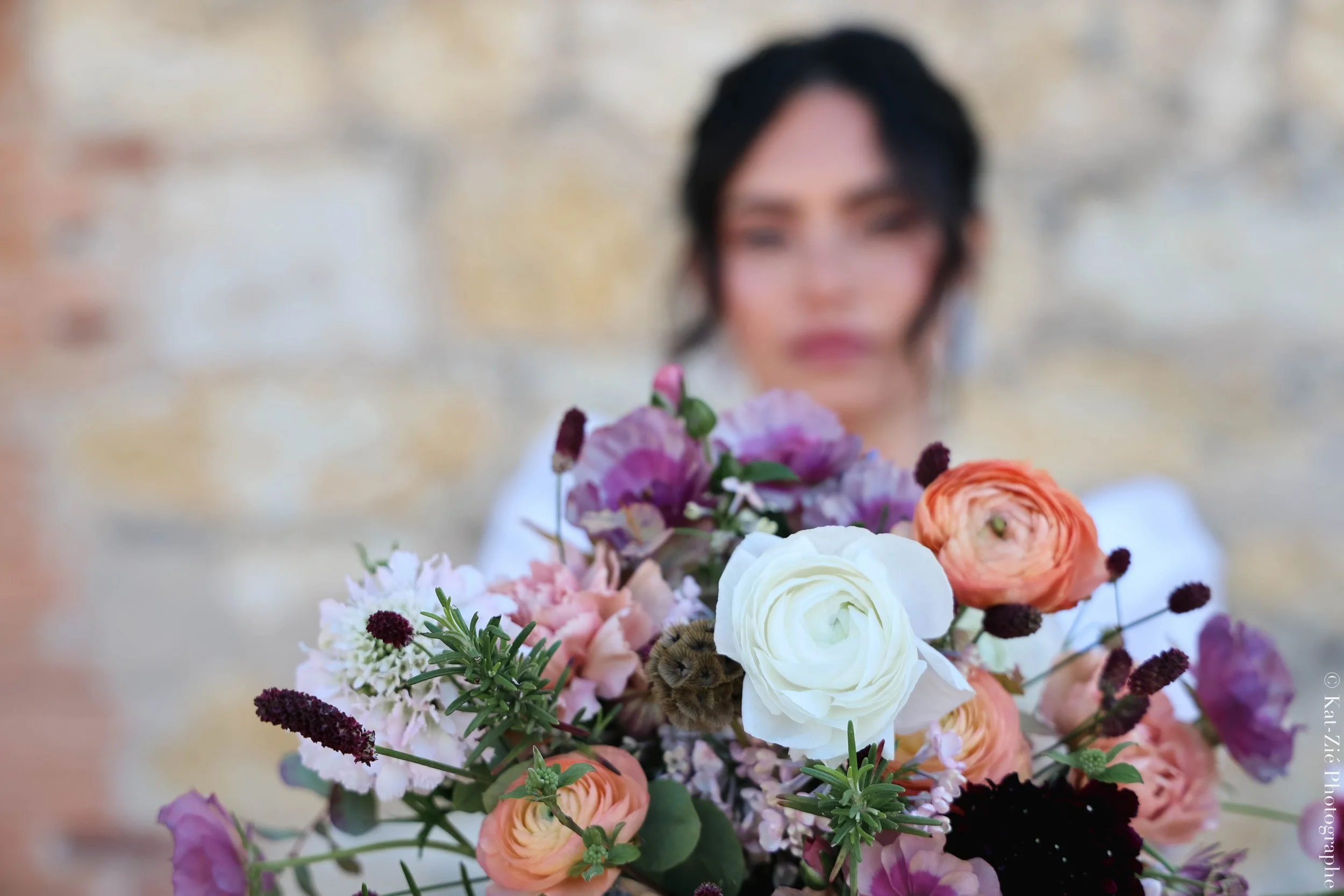 Une femme avec des cheveux noirs et une chemise blanche, tenant un bouquet de fleurs colorées devant elle, avec un mur en pierre en arrière-plan.