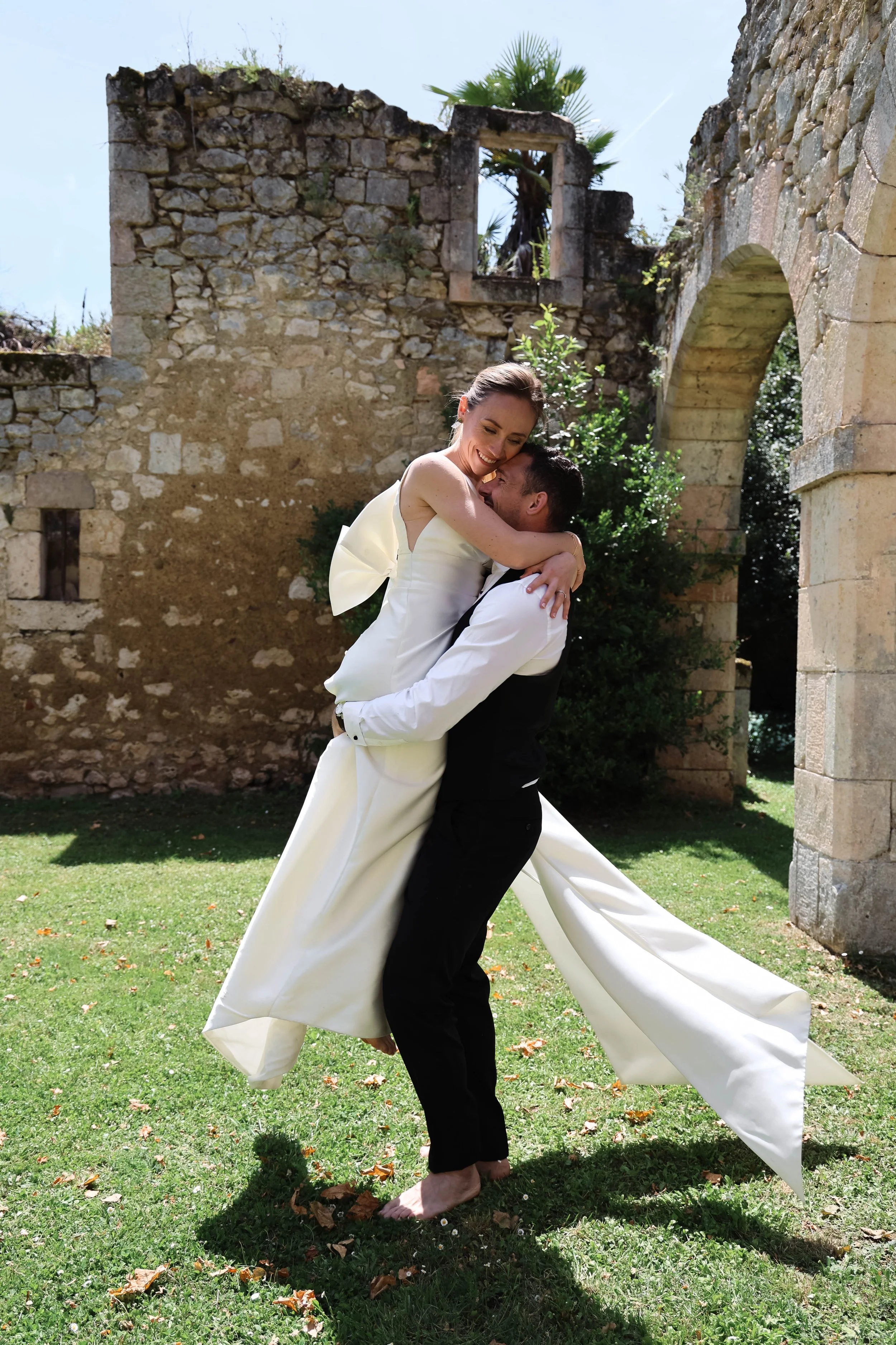Un couple de mariés dansants, la femme en robe blanche et le homme en costume noir, en extérieur près de ruines anciennes, sous un ciel ensoleillé.