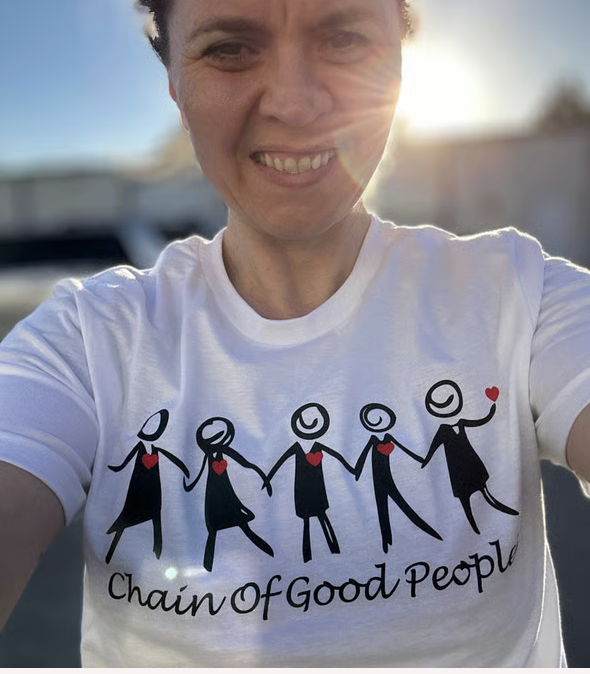 Person taking a selfie outdoors wearing a white T-shirt with a graphic of people holding hands and the text 'Chain of Good People'.