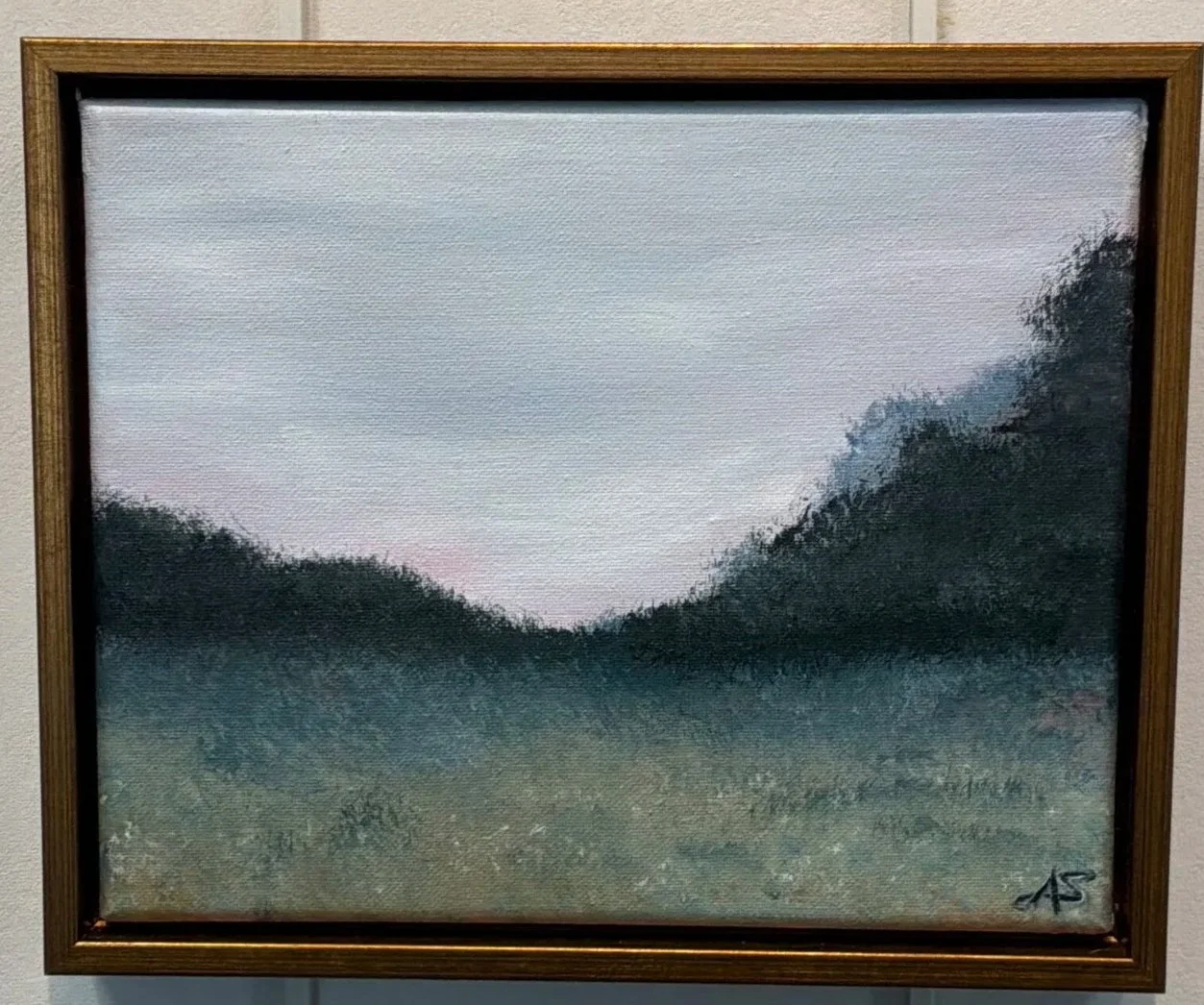 A painting of a natural landscape with a cloudy sky, tree-covered hills, and grassy foreground, framed in wood.