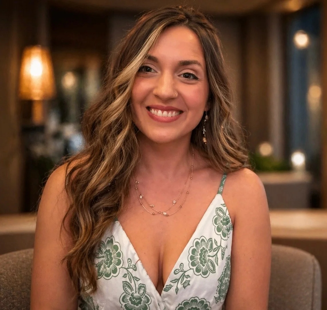 A smiling woman with long, wavy brown hair sitting indoors, wearing a floral dress, jewelry, and a nose ring.