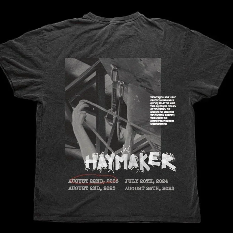 Black T-shirt featuring a graphic of a person doing a pull-up on gym rings, with the word 'HAYMAKER' in bold graffiti style text below. The shirt also displays several dates and a paragraph of small text about the belay responsibilities