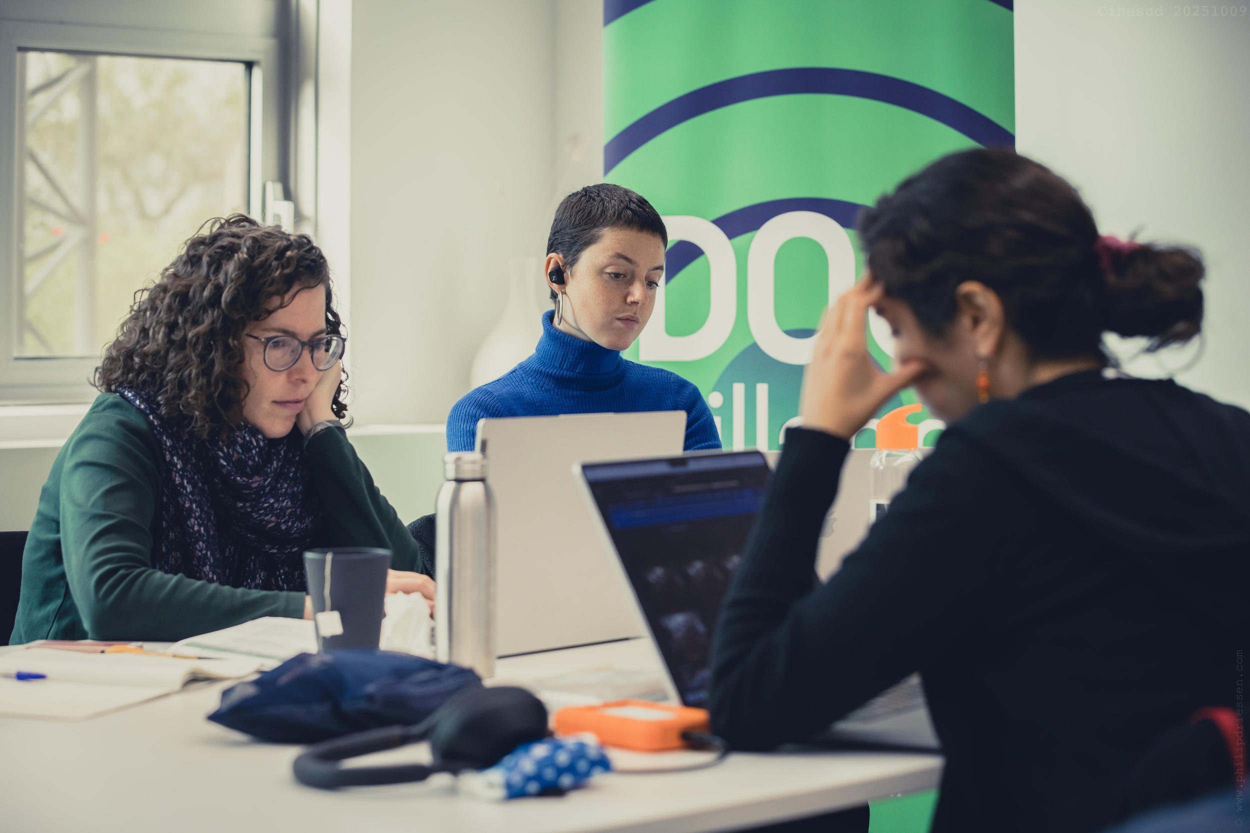 Tre kvinder, som sidder ved et bord med deres bærbare computere og koncentrerer sig om, hvad de ser på skærmen. Bag dem er et skilt, hvor der står "DOCvillage"