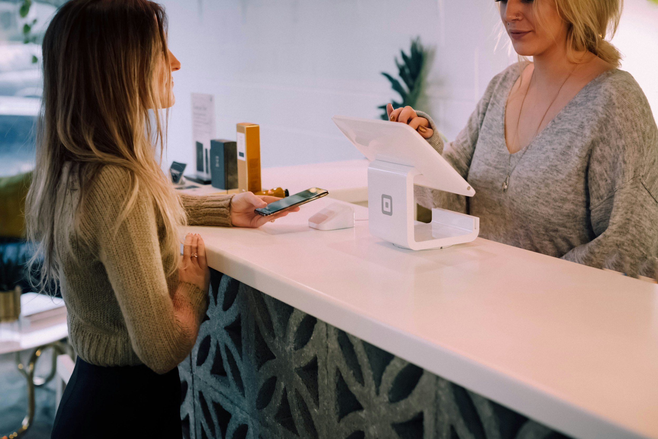 A woman at a reception desk purchases something using her smartphone while a cashier or receptionist processes the transaction with a tablet at a modern counter.