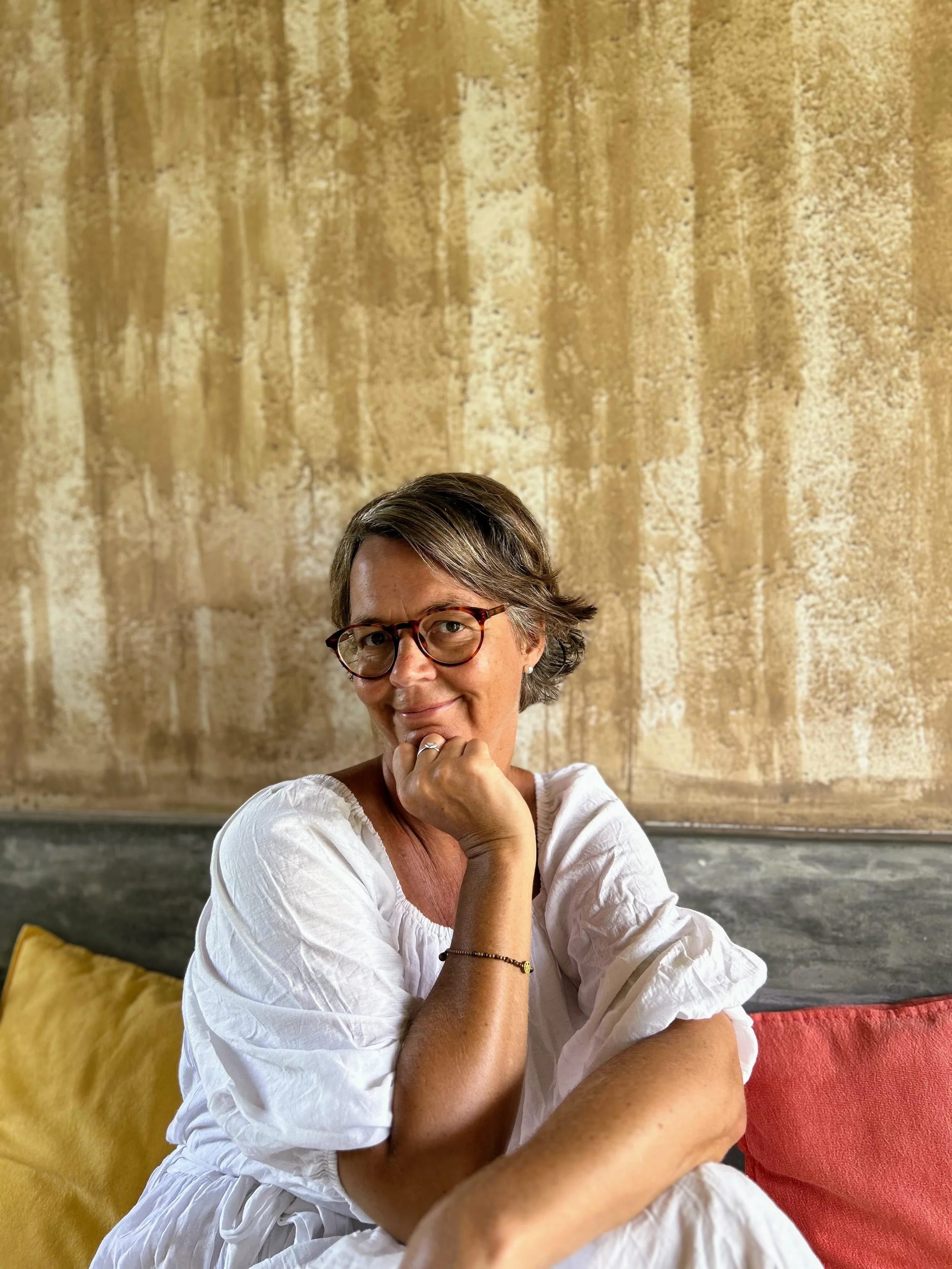 Eine Frau sitzt entspannt auf einem Sofa, trägt eine weiße Bluse, hat kurze braune Haare und trägt randlose Brille, vor einem goldenen Hintergrund.