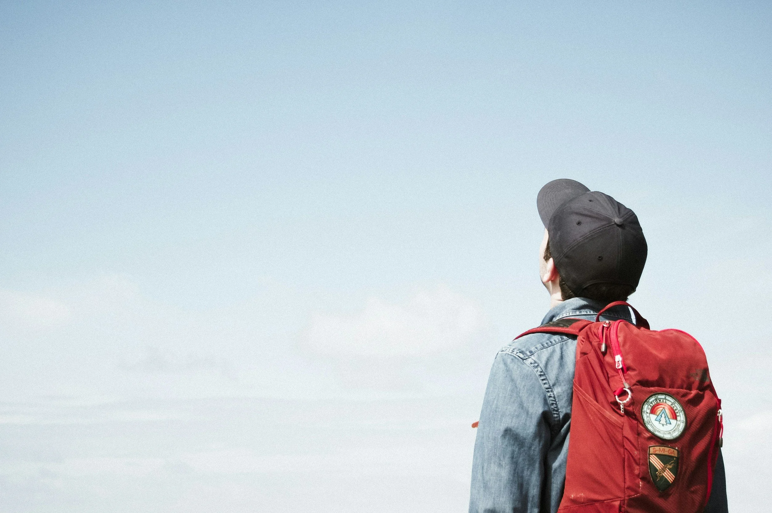 Person wearing a black cap and denim jacket, with a red backpack, looking up at the sky.
